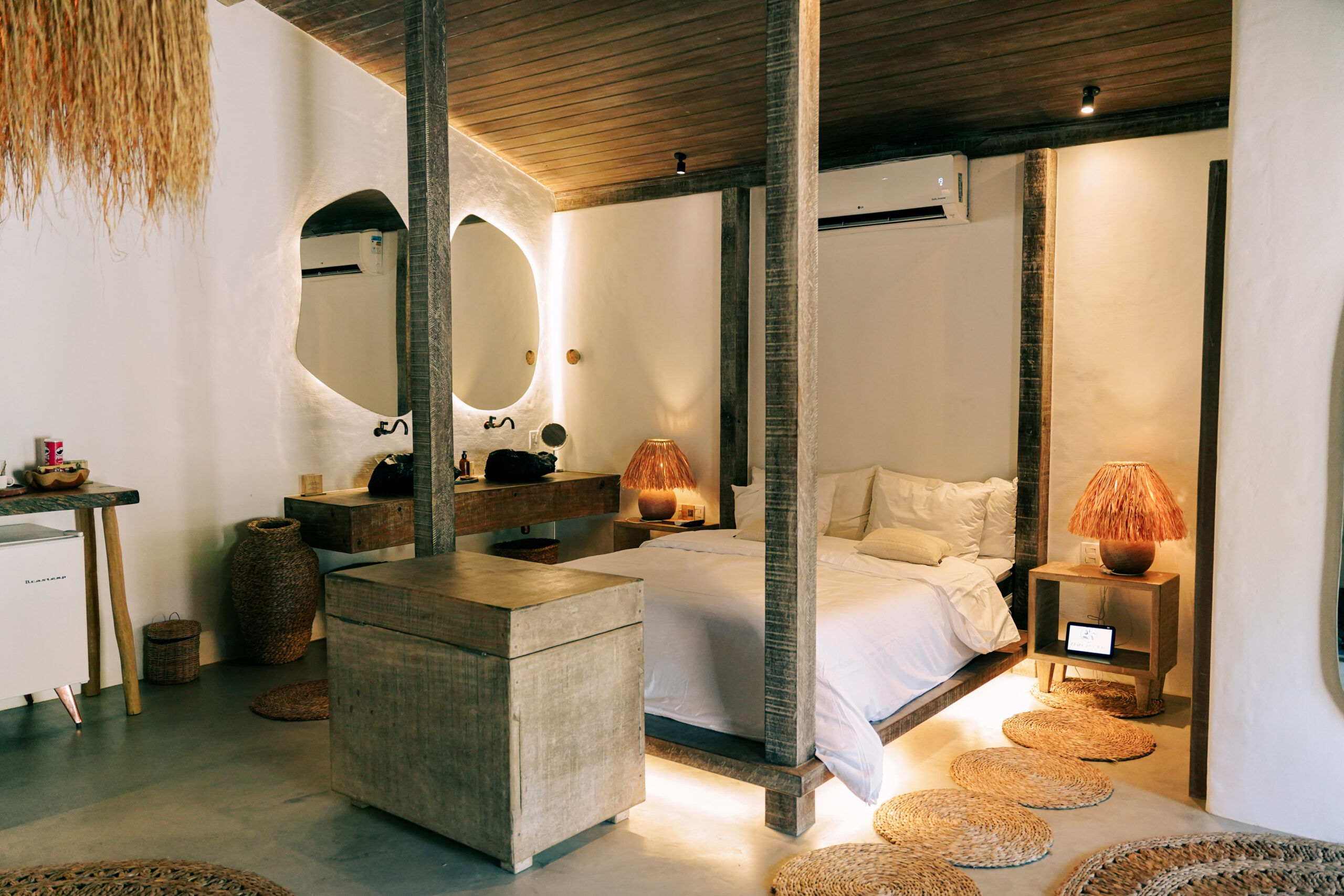 Interior of a bedroom – a double bed with white sheets and grey-wood four poster beams, a grey-wood cabinet at the foot of the bed, rugs on the floor, white walls and an odd-shaped mirror on the far wall with two sinks carved out of dark rocks.