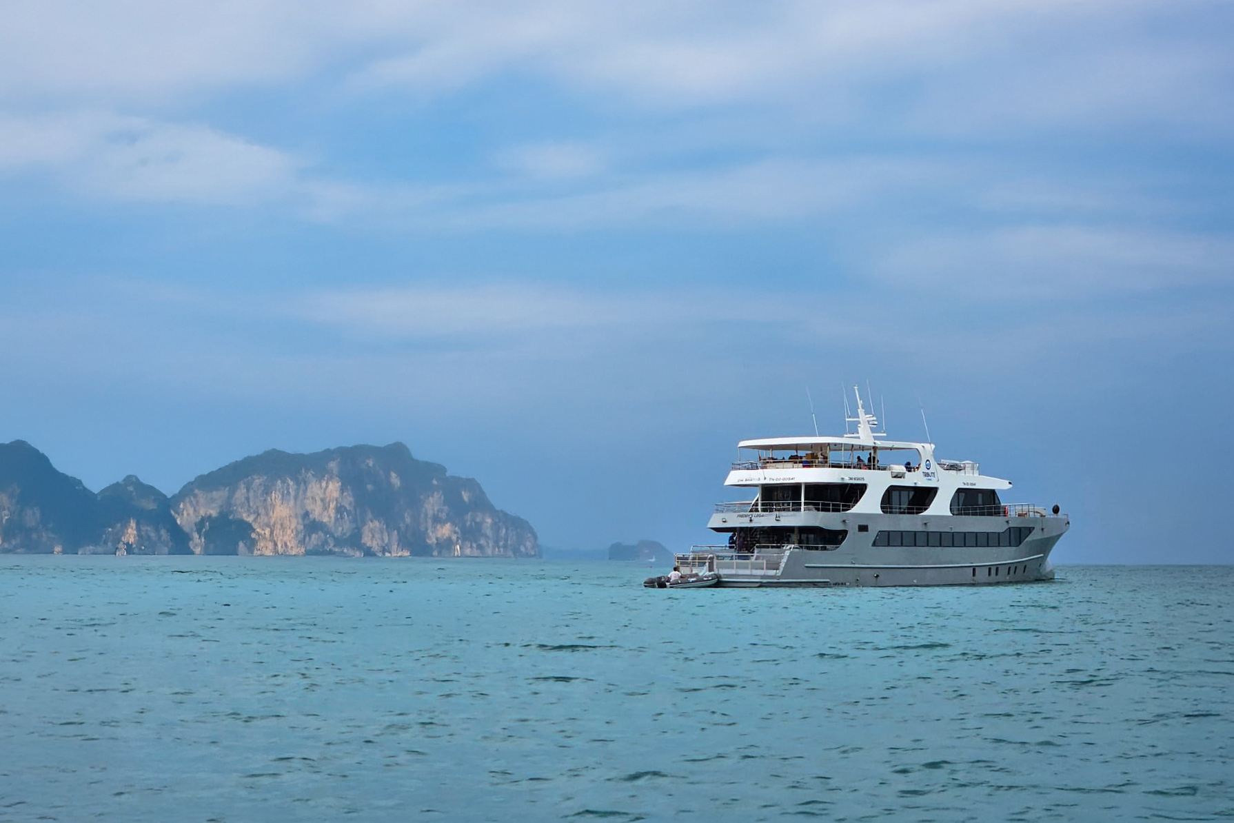 Yacht sailing on turquoise blue water with mountain cliffs and blue sky in the background.