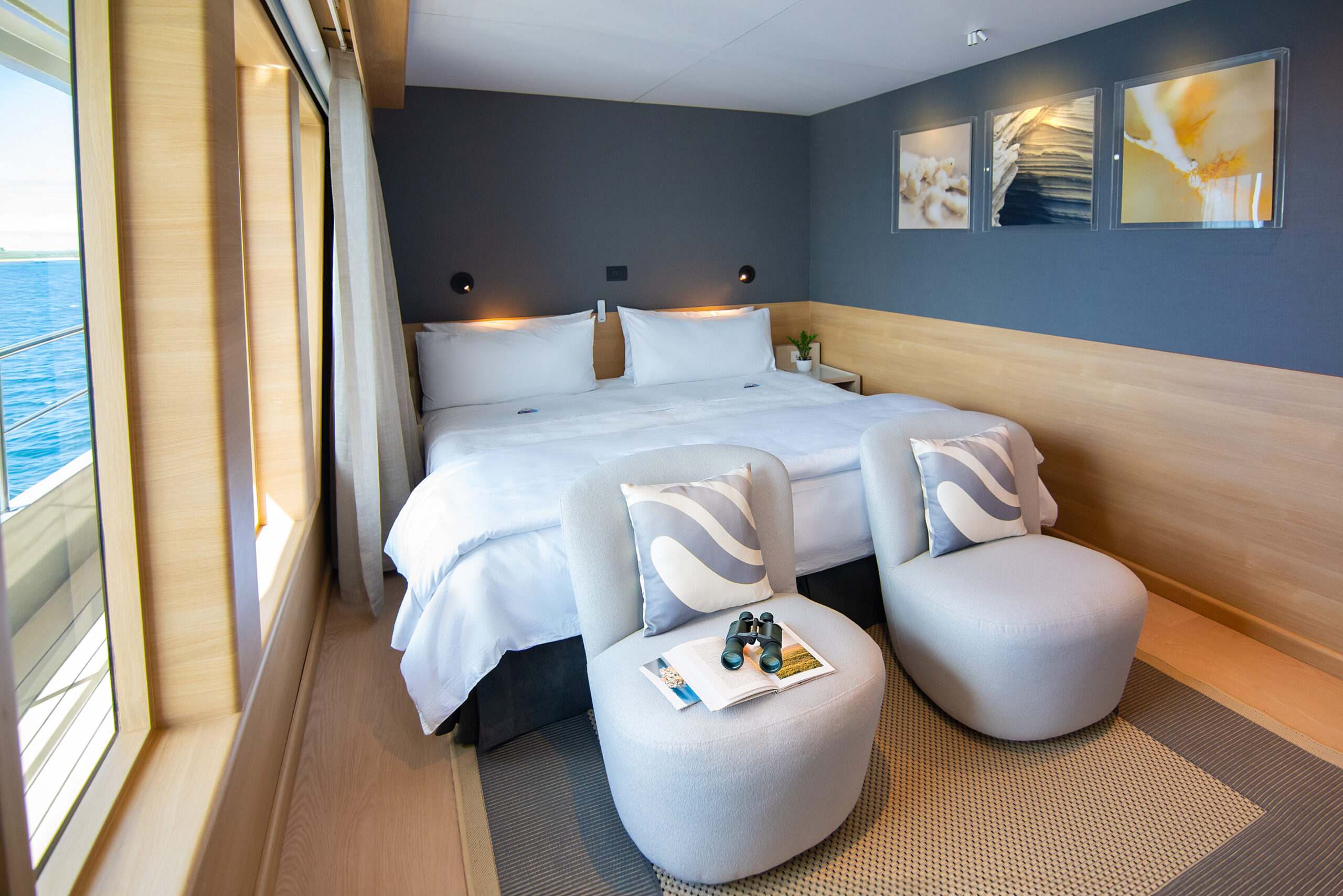 Double bed with white sheets and footstools at the base in a carpeted room with grey walls and paintings on one wall.