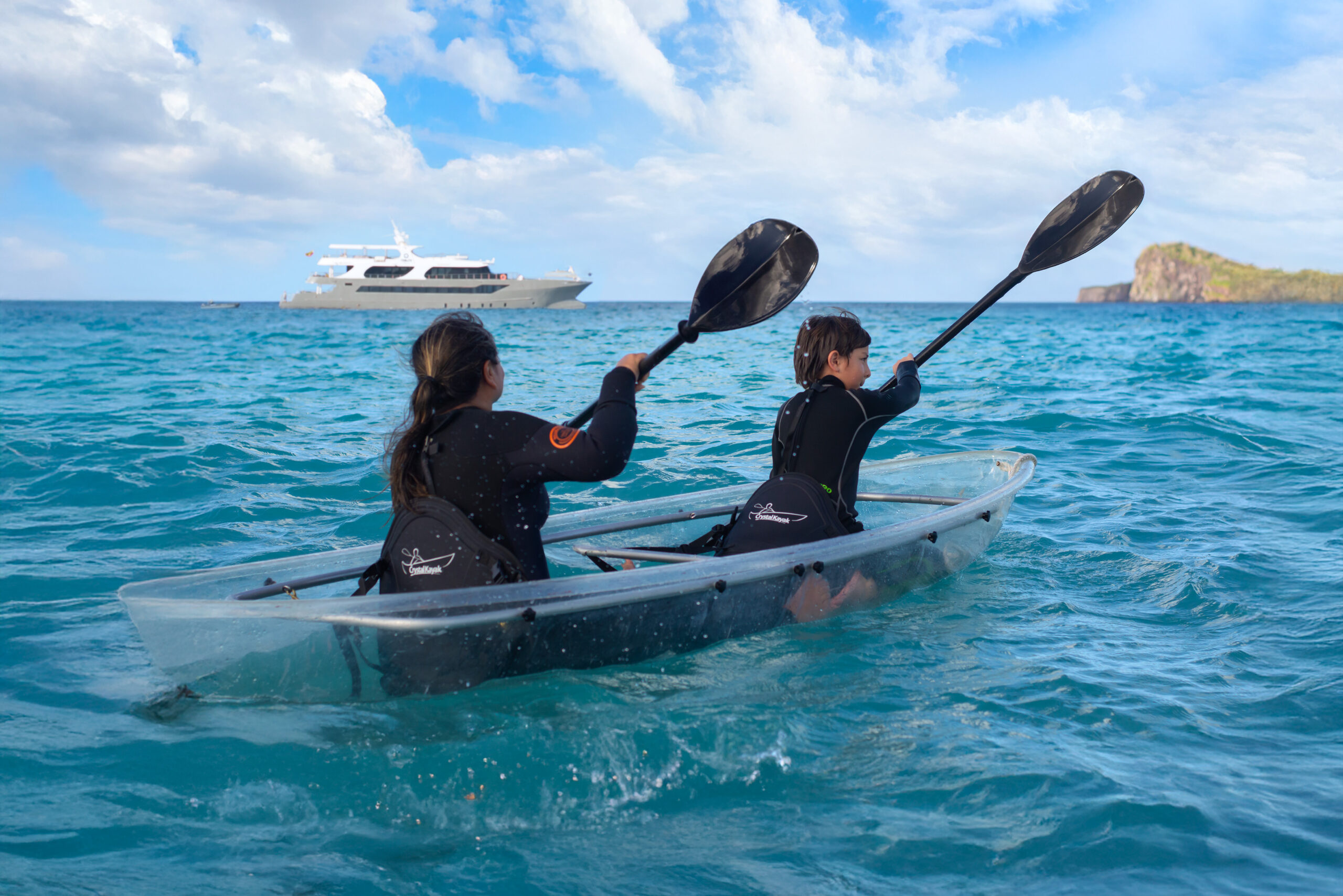 Two people in black wetsuits paddling a kayak on open blue ocean with a yacht in the far distance.