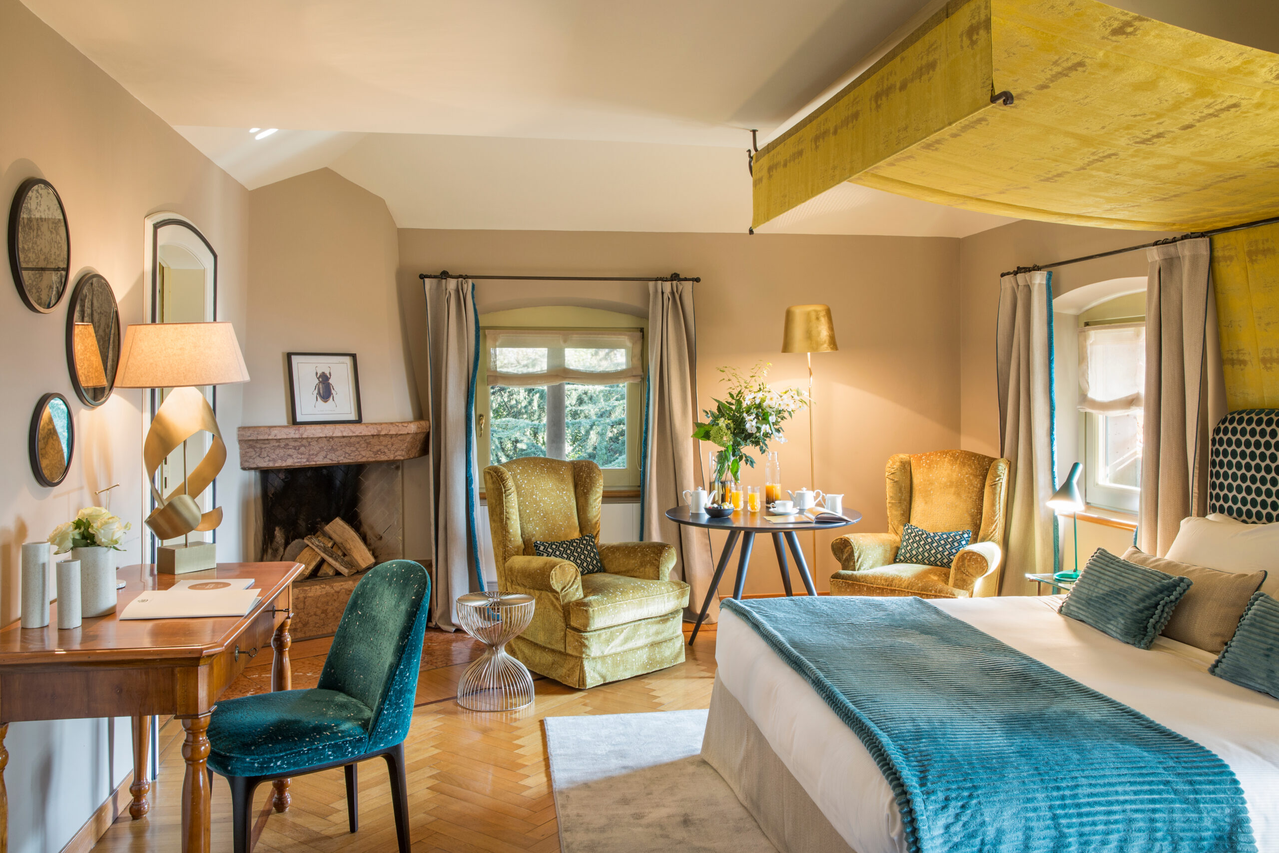 Bedroom scene showing a double bed with white and blue sheets, a desk with blue chair, and a green plush lounge chair, in a room with tan carpet, warm lighting and a lot of mirrors on the wall.