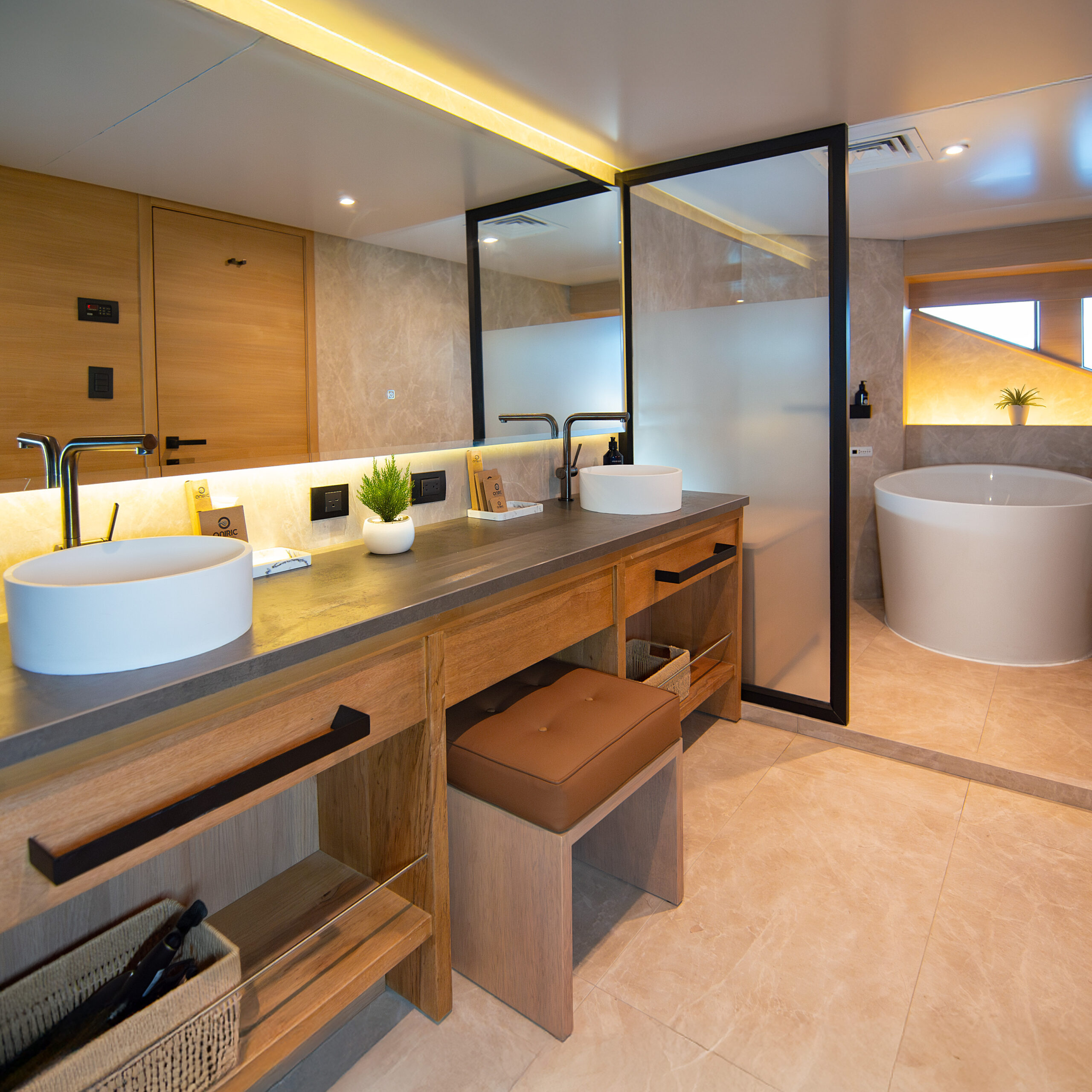 Bathroom with round white sink, grey countertops, a big mirror, light wood drawers and in the background a shower and round bathtub.