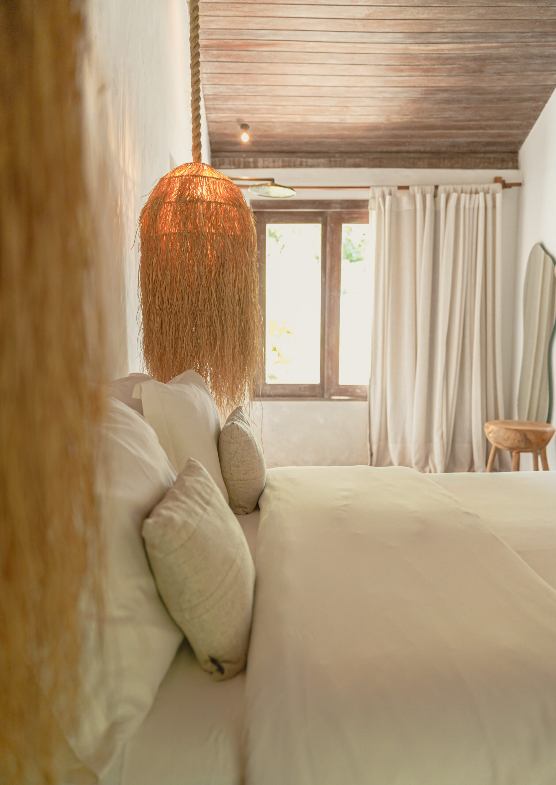 Side view of a bed with white and beige sheets, looking over the bed toward a bright window. On each side of the bed are two hanging lamps with dried grass for lamp shades.