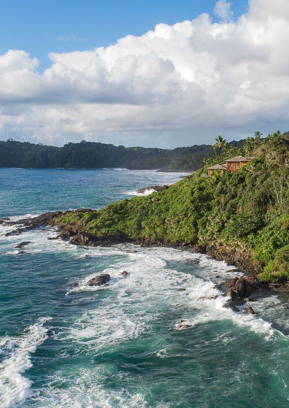 Distant view of a villa with pitched roof and wrap-around terraces set in thick palm jungle overlooking a rocky coastline with blue ocean and waves crashing.
