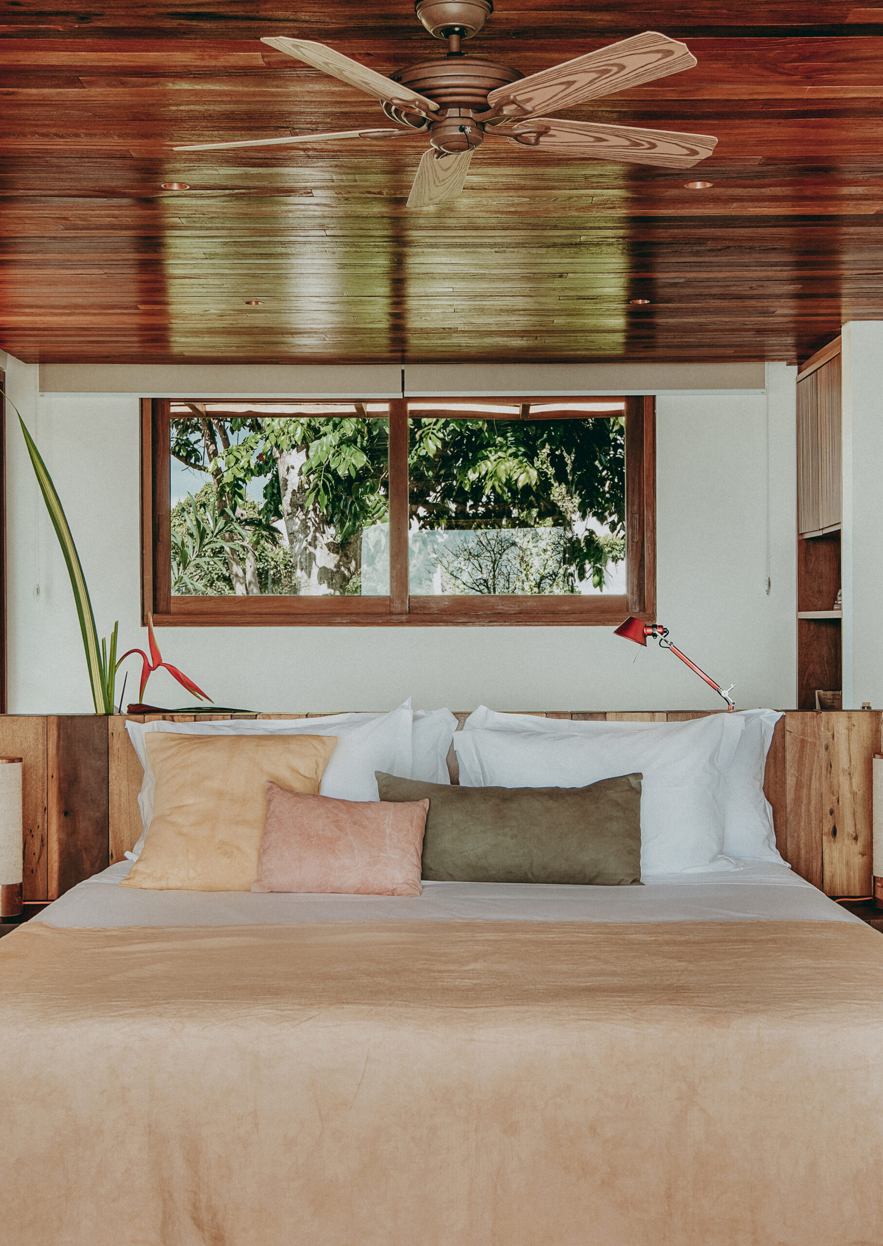 Interior of a double bed with beige, green and white sheets and pillows, with a small rectangular window and ceiling fan above, and redwood ceiling.