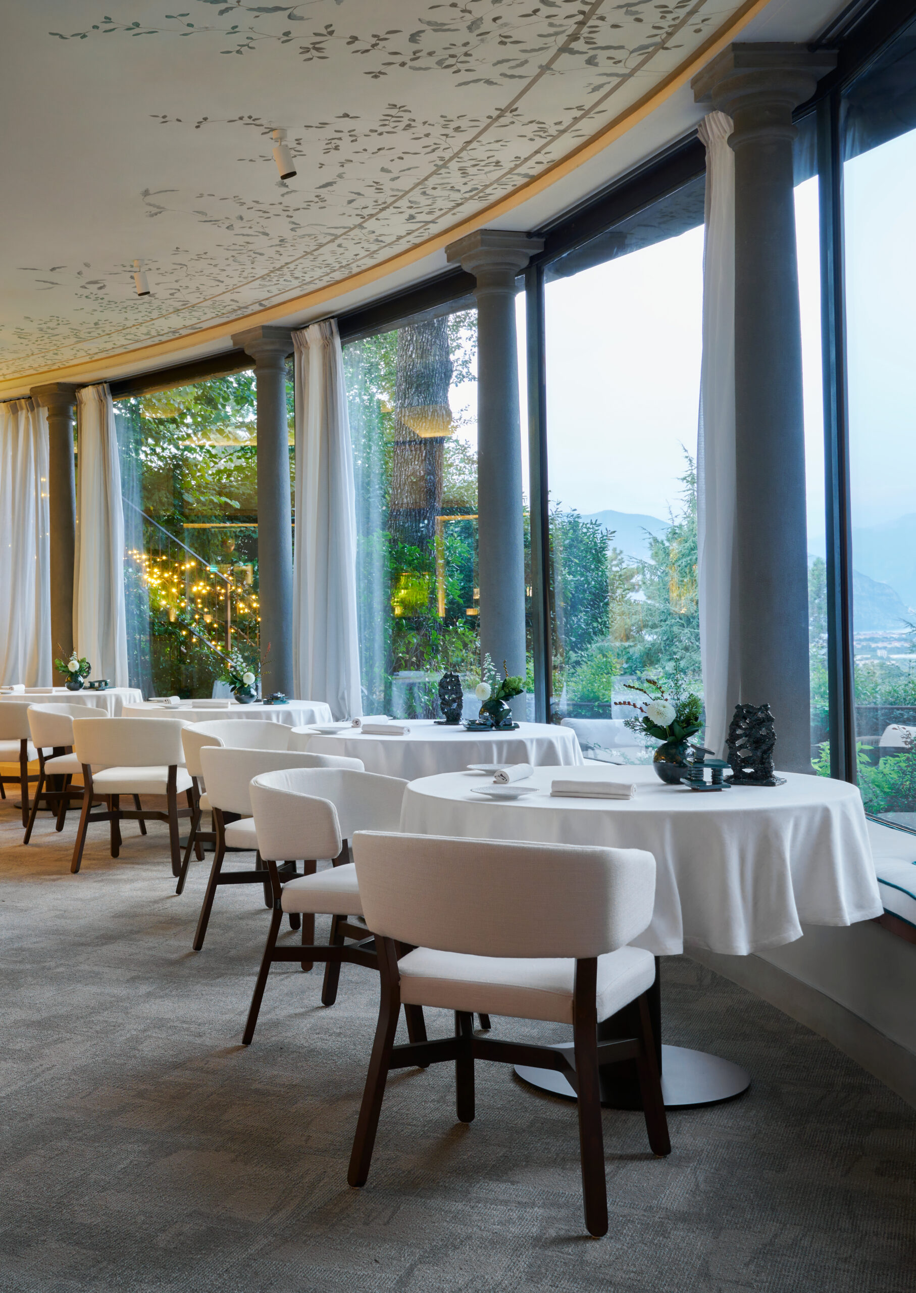 Several formal tables dressed in white tablecloths with white dining chairs in a grey carpeted room with large floor-to-ceiling picture windows overlooking a nature scene.
