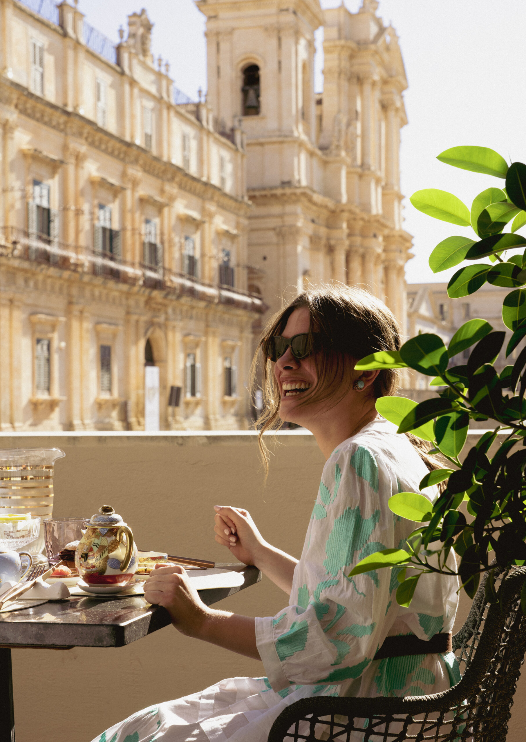 A woman in a white outfit sitting at a chair on an outdoor terrace smiling, with Baroque buildings in the background.