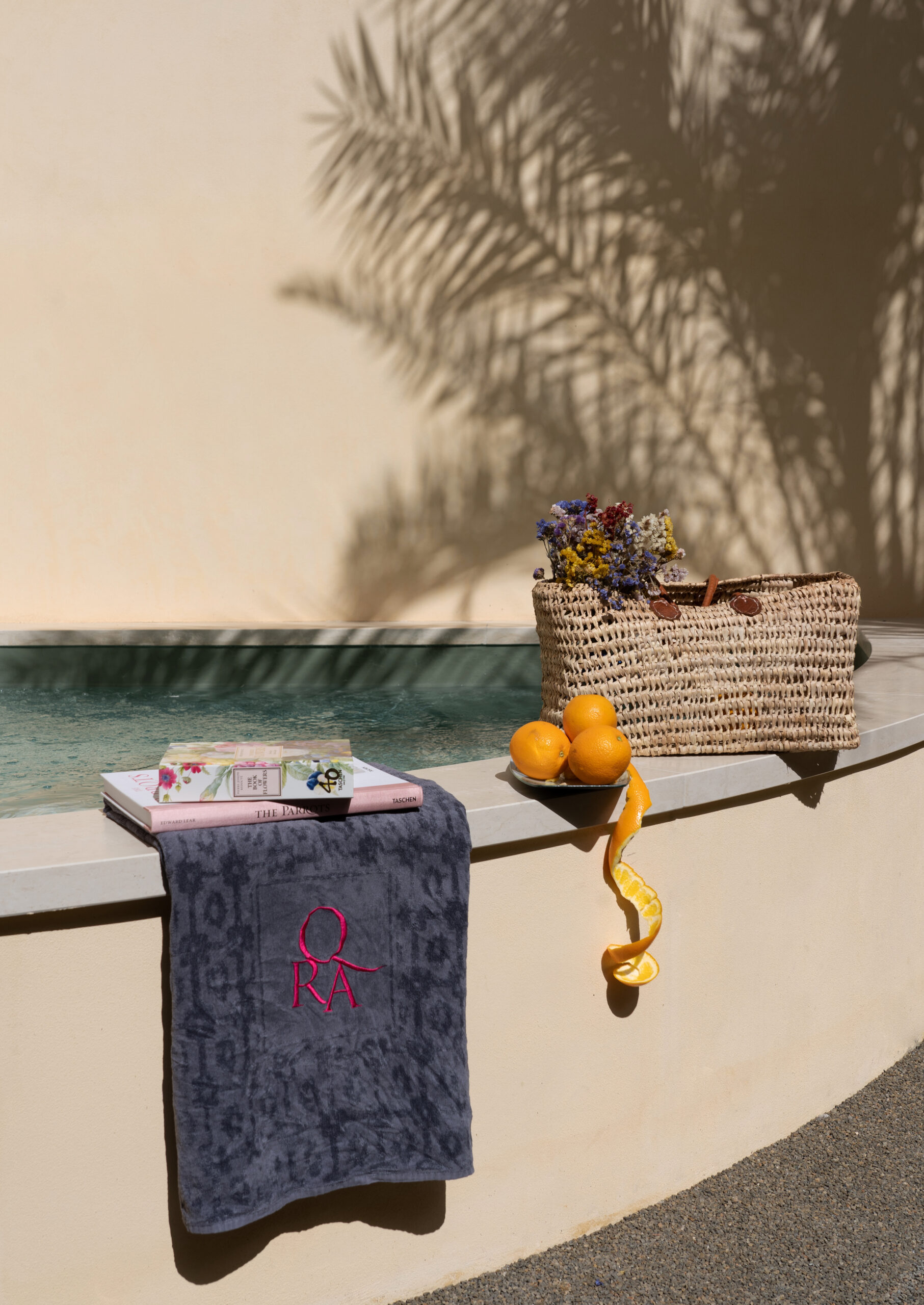 Exterior of the ledge of a pool with a towel, orranges and snacks, and the shadow of a palm tree.
