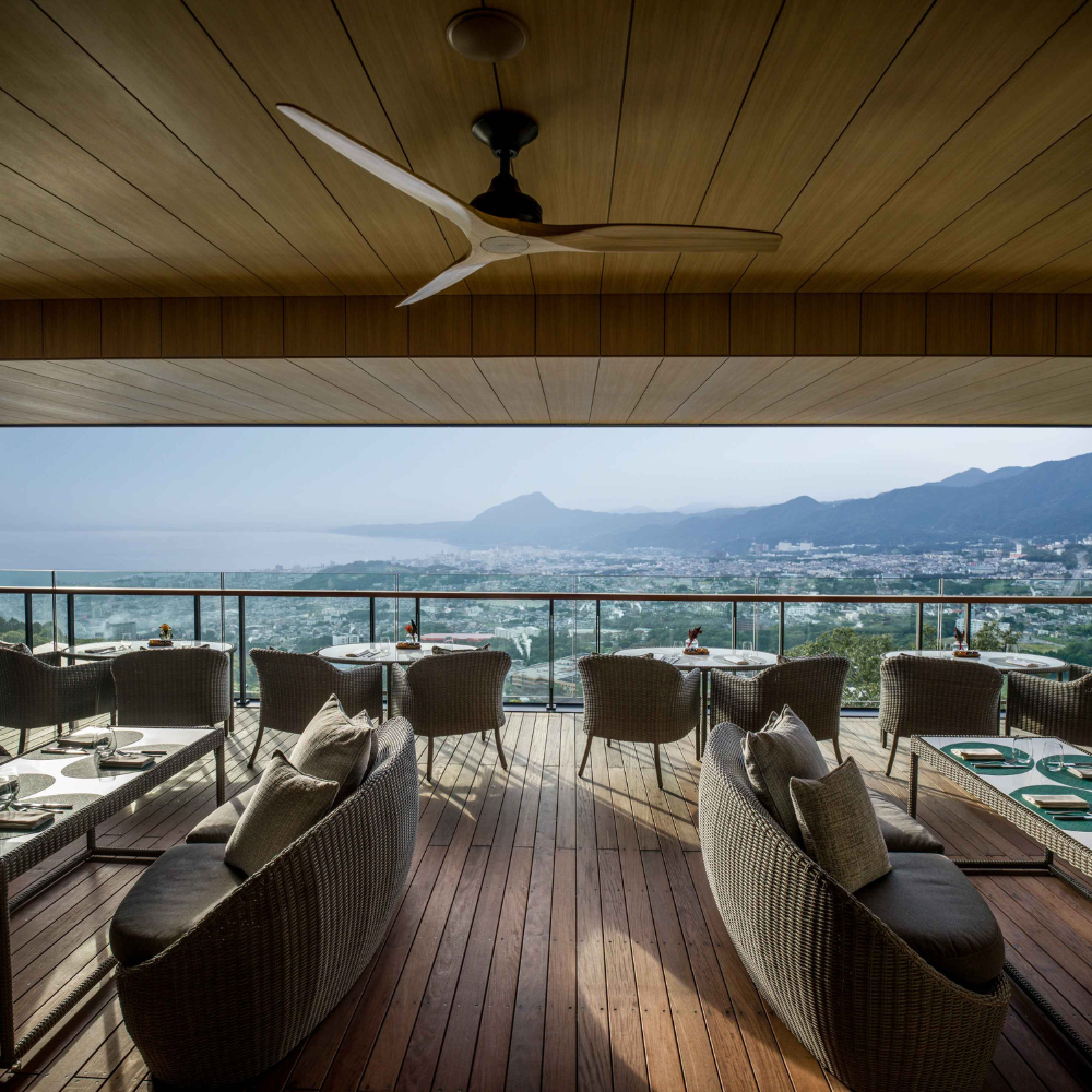 A covered terrace with tables and chairs and a panoramic view of the mountains and sea