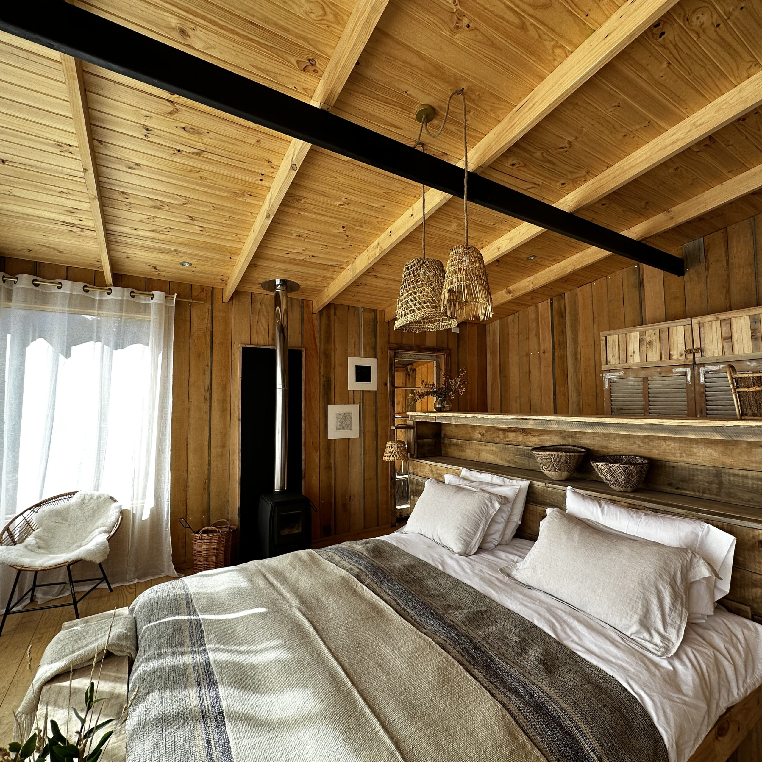Bedroom with a wooden ceiling and walls, a large bed with neutral linens, woven pendant lights, a wood burner and sheer curtains filtering natural light.