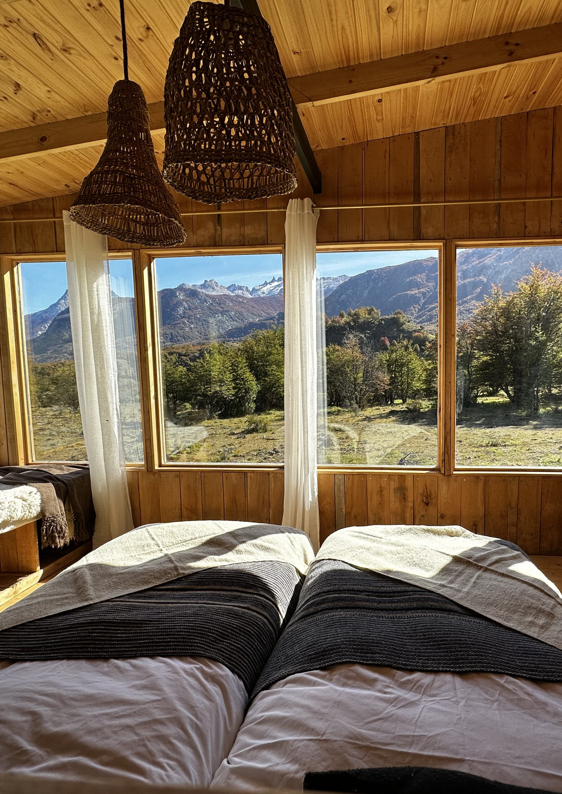 Twin beds with woven blankets positioned before floor-to-ceiling windows overlooking a landscape of trees and rugged Patagonian mountains.