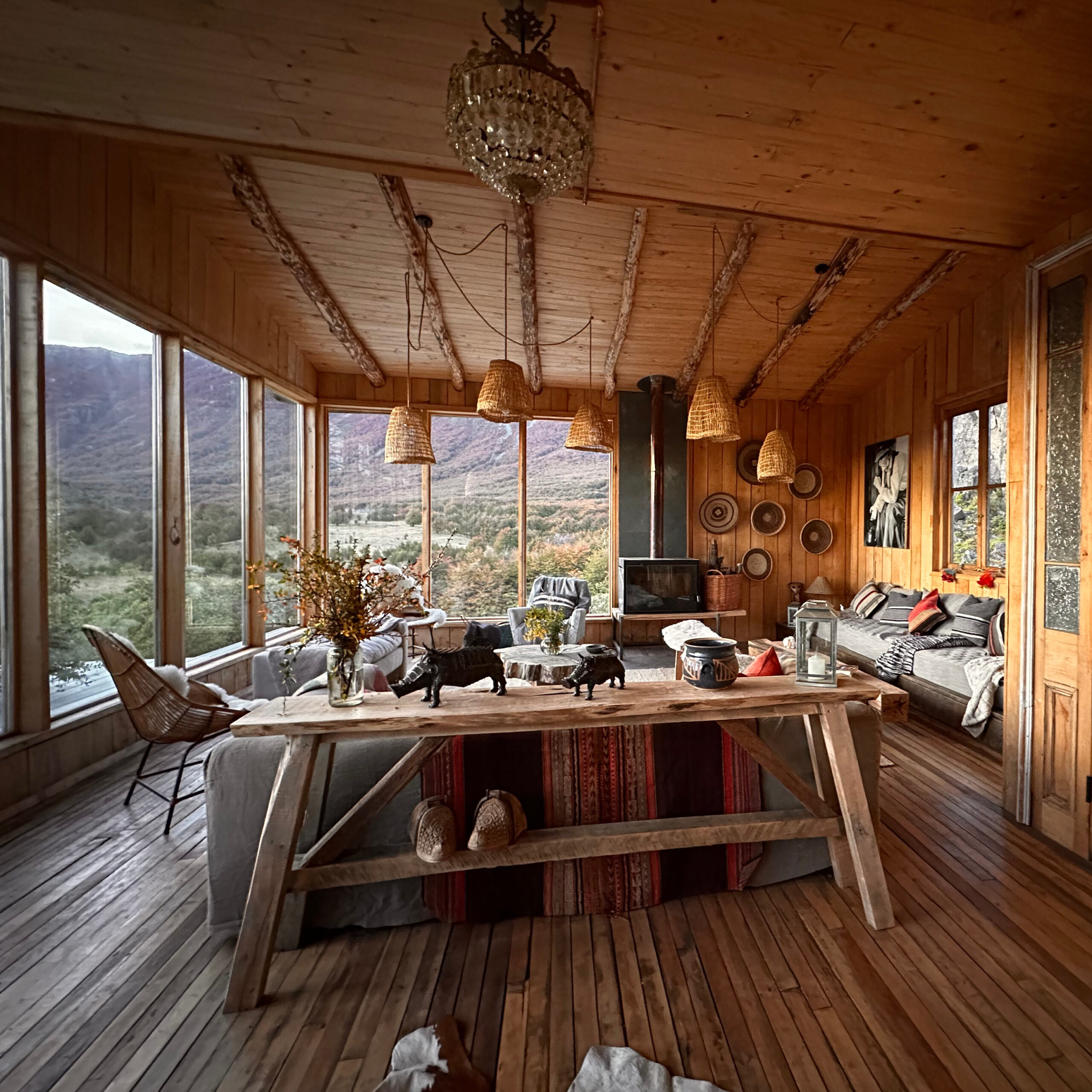 Open living and dining space with wide windows overlooking Patagonian scenery, wooden floors, wicker pendant lights, cushioned sofas and a long wooden table with decorative objects.