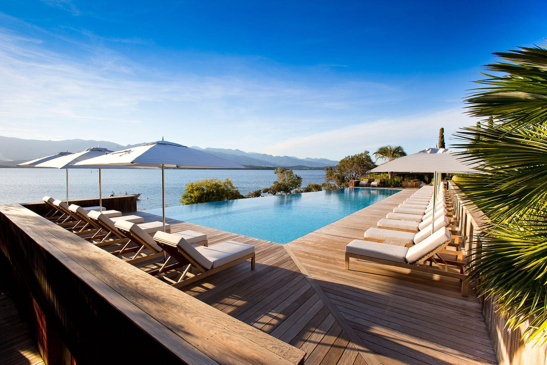 Pool with blue water, sun loungers and umbrellas along a wooden deck, and in the background it overlooks the sea.
