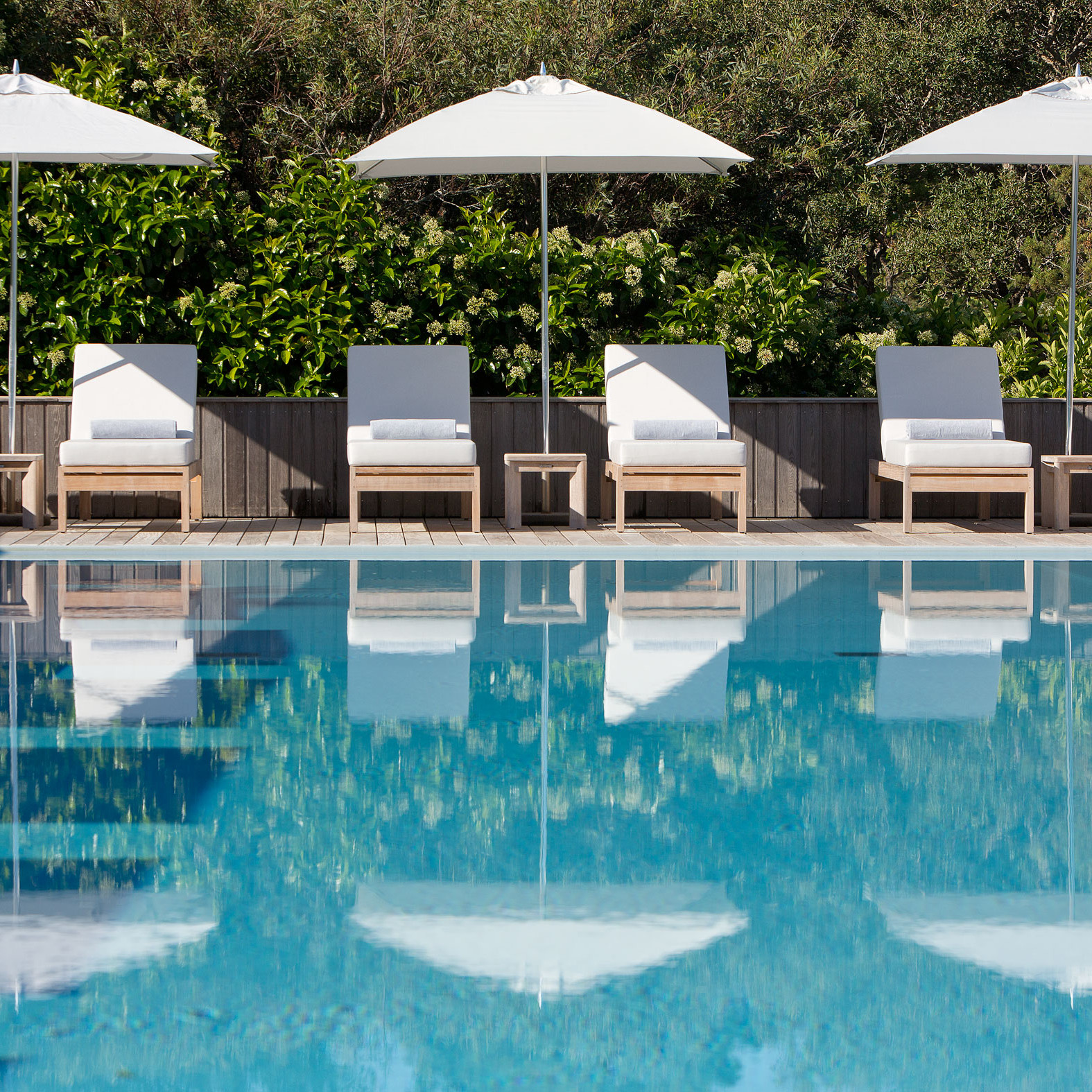 Blue pool water in foreground with four white sun loungers and umbrellas in the background.
