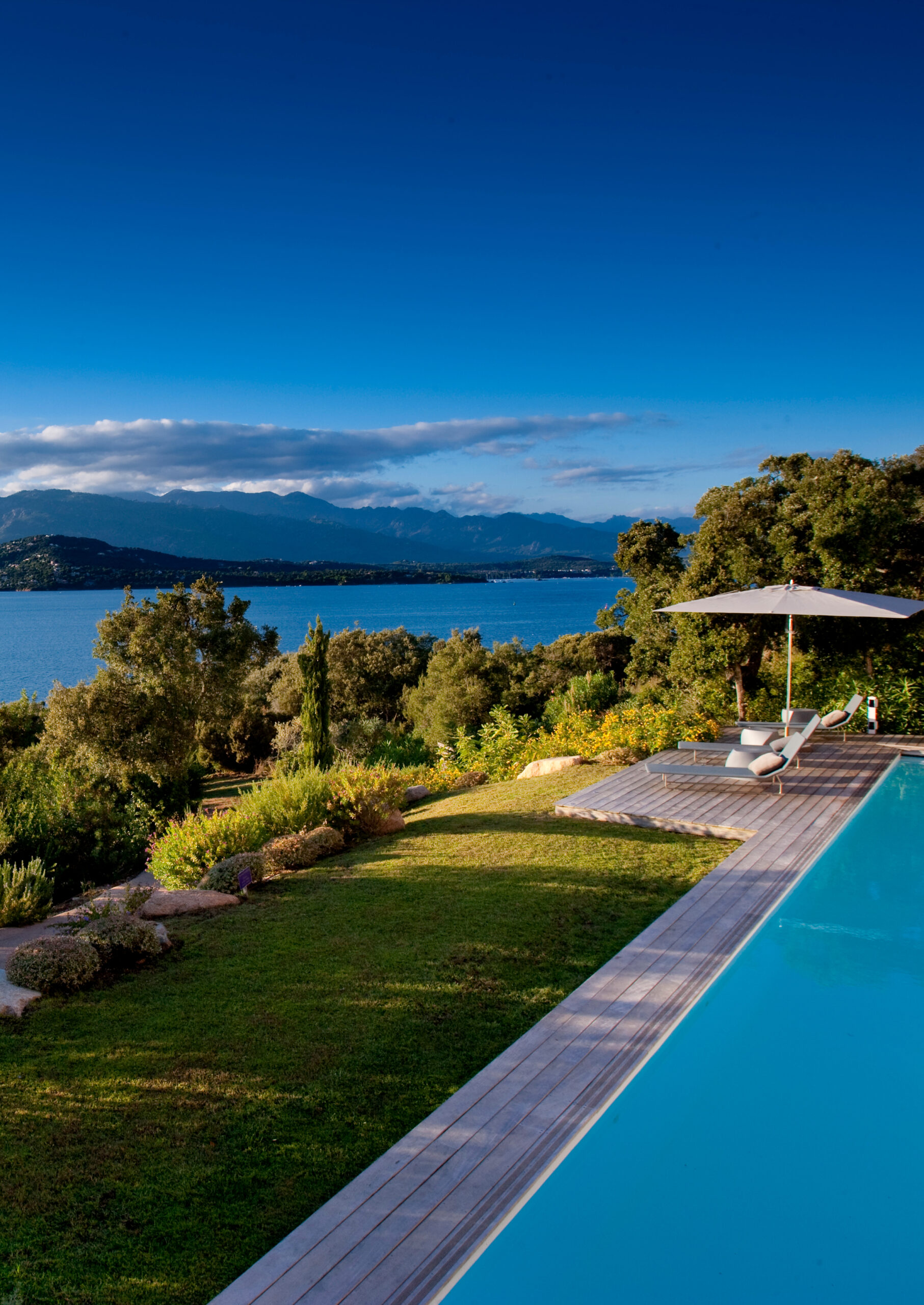Pool with blue water, a sun lounger and umbrella along a wooden deck, green lawns and trees, and in the background it overlooks the sea.