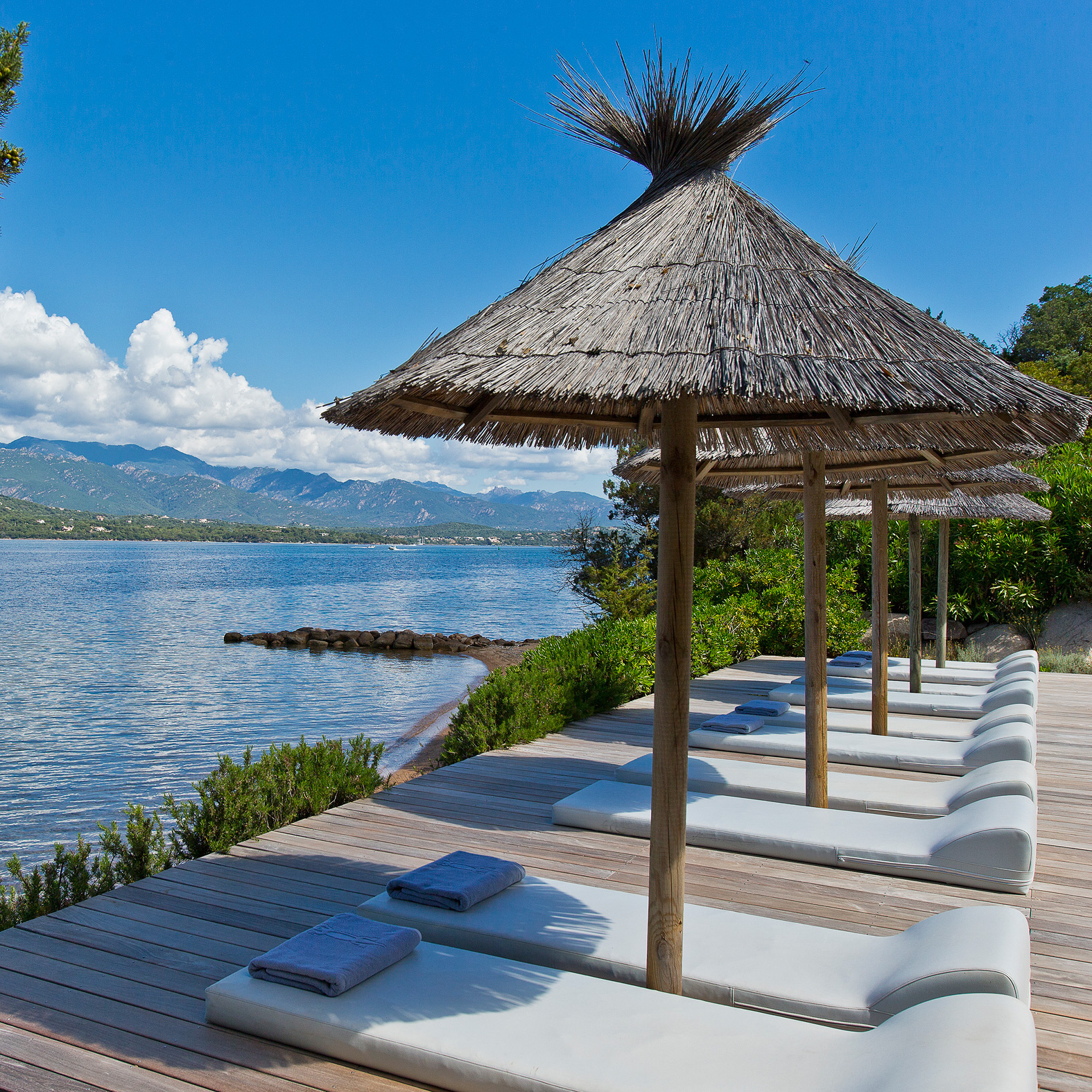 Row of white sun loungers and thatched umbrellas set up overlooking the sea and mountains beyond.