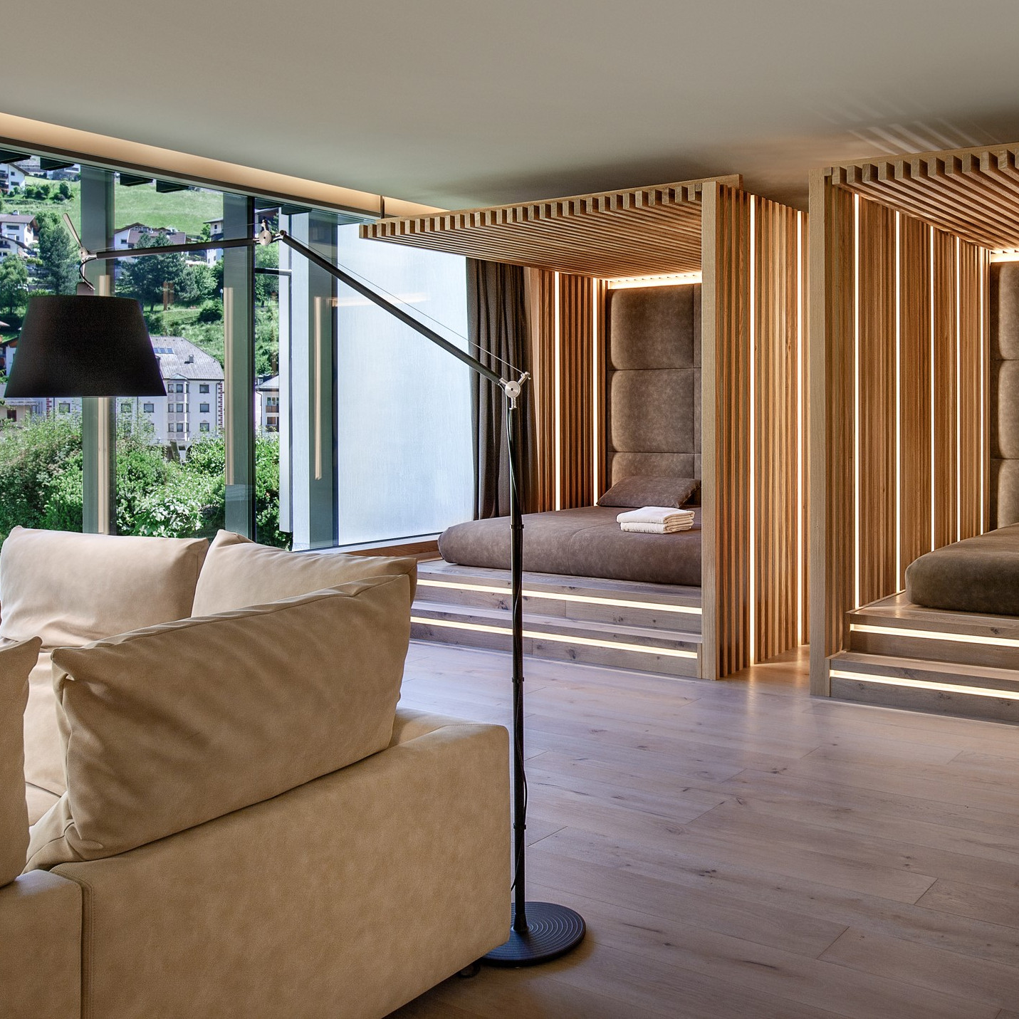 Interior of a room with Japanese-style futon beds with brown bedding and wooden interior covers. In the foreground to the left is a tan sofa and a large black lamp, and in the background is a window with a green mountainside and buildings.