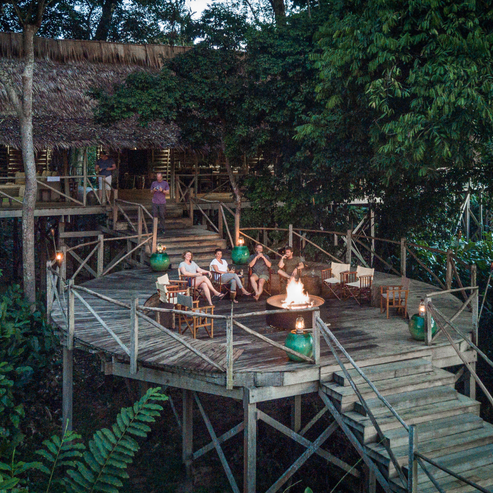Four smiling people sitting in chairs on a raised round wooden deck with a bonfire