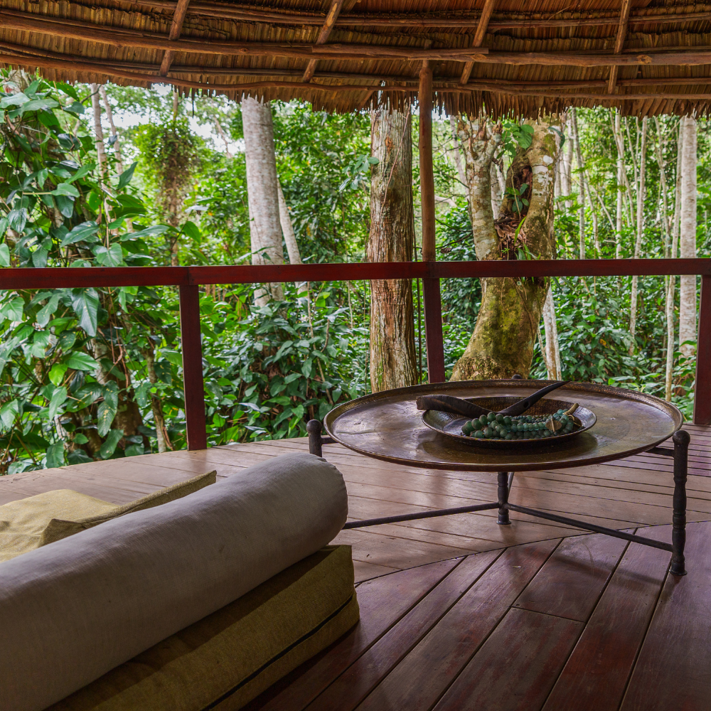 A covered deck with a round table, the back of a couch and views of green foliage