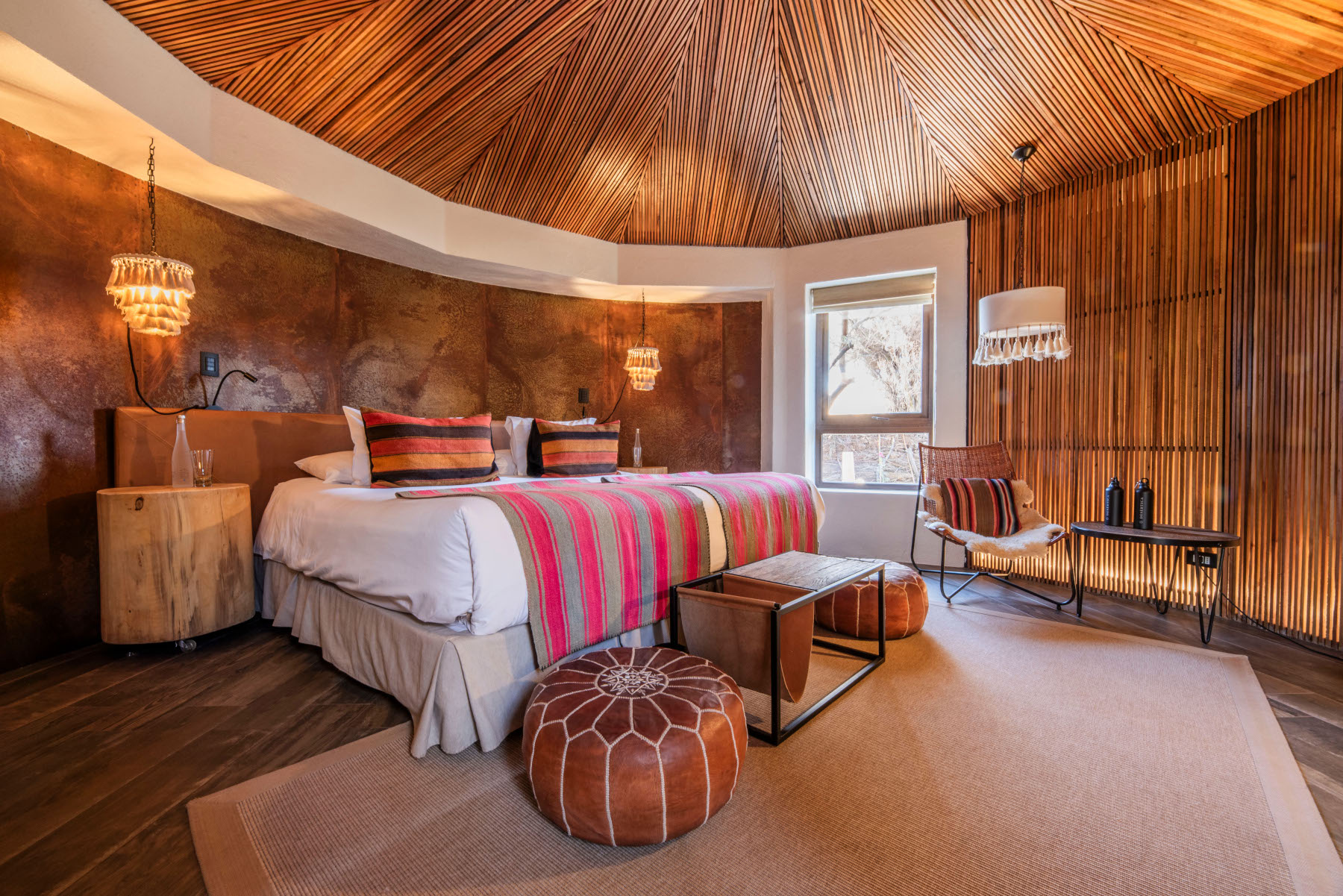 Interior of a round bedroom with wooden or thatch walls, a bed with grey and colourful pink textile bedding, and lampshades and footstools in textiles and leather.