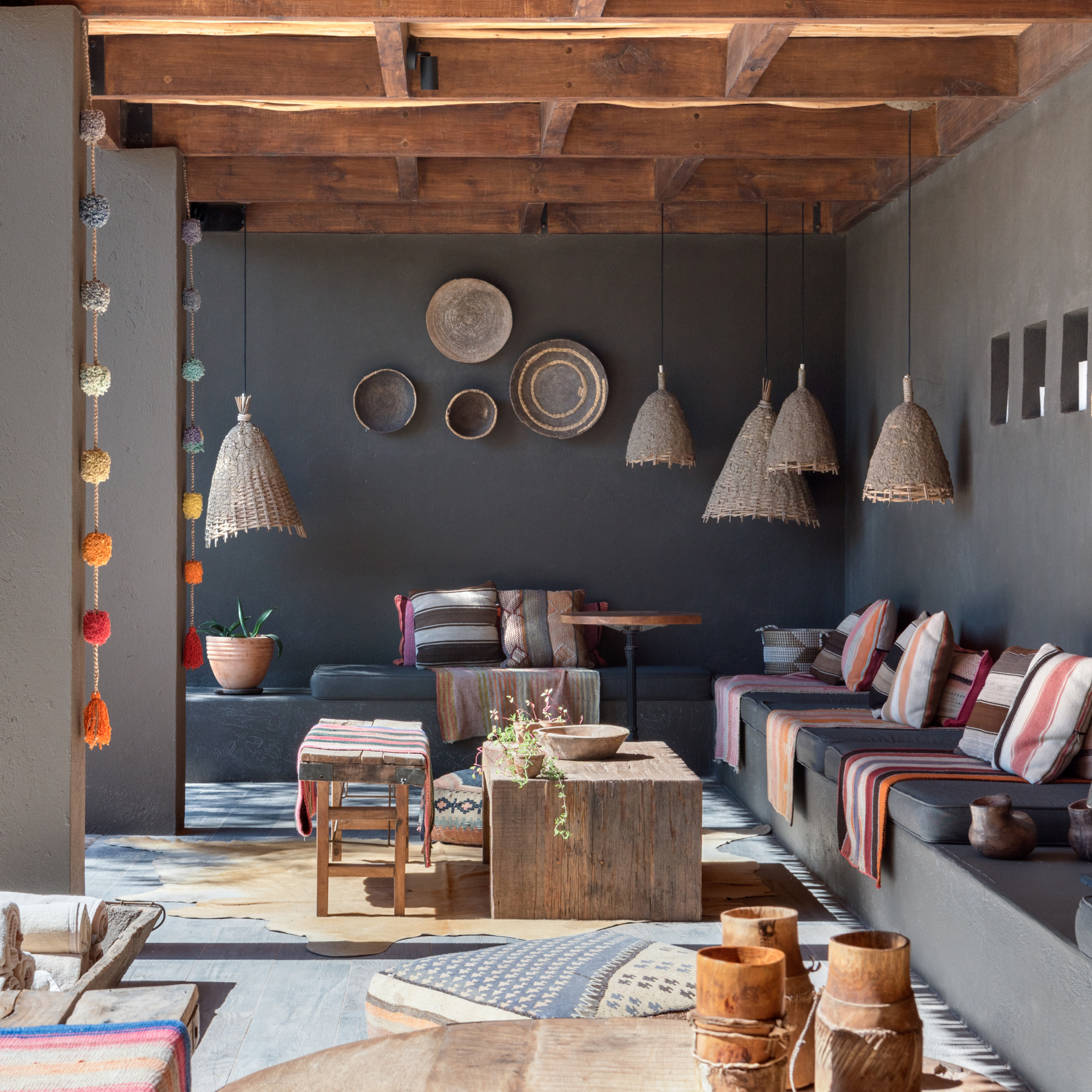 Exterior of a shaded seating area with grey walls and a long banquette on the right with multi-coloured woven pillows and blankets. On the walls are baskets and overhead are basket lampshades. The sun shines in on part of the terrace where there are low tables and stools.