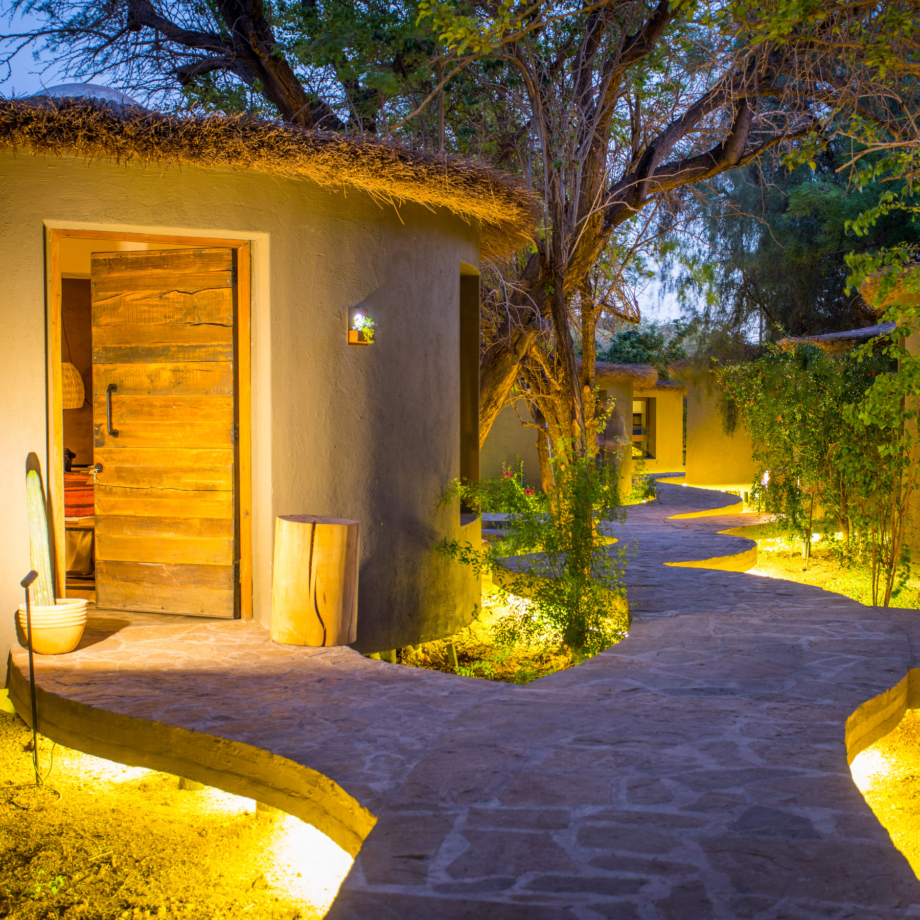 Evening shot of a paved stone walkway leading to a room with thatched roof and wooden door slightly ajar. Alongside the footpath are green trees and plants and low lighting below the stone lighting up the scene with yellow light.