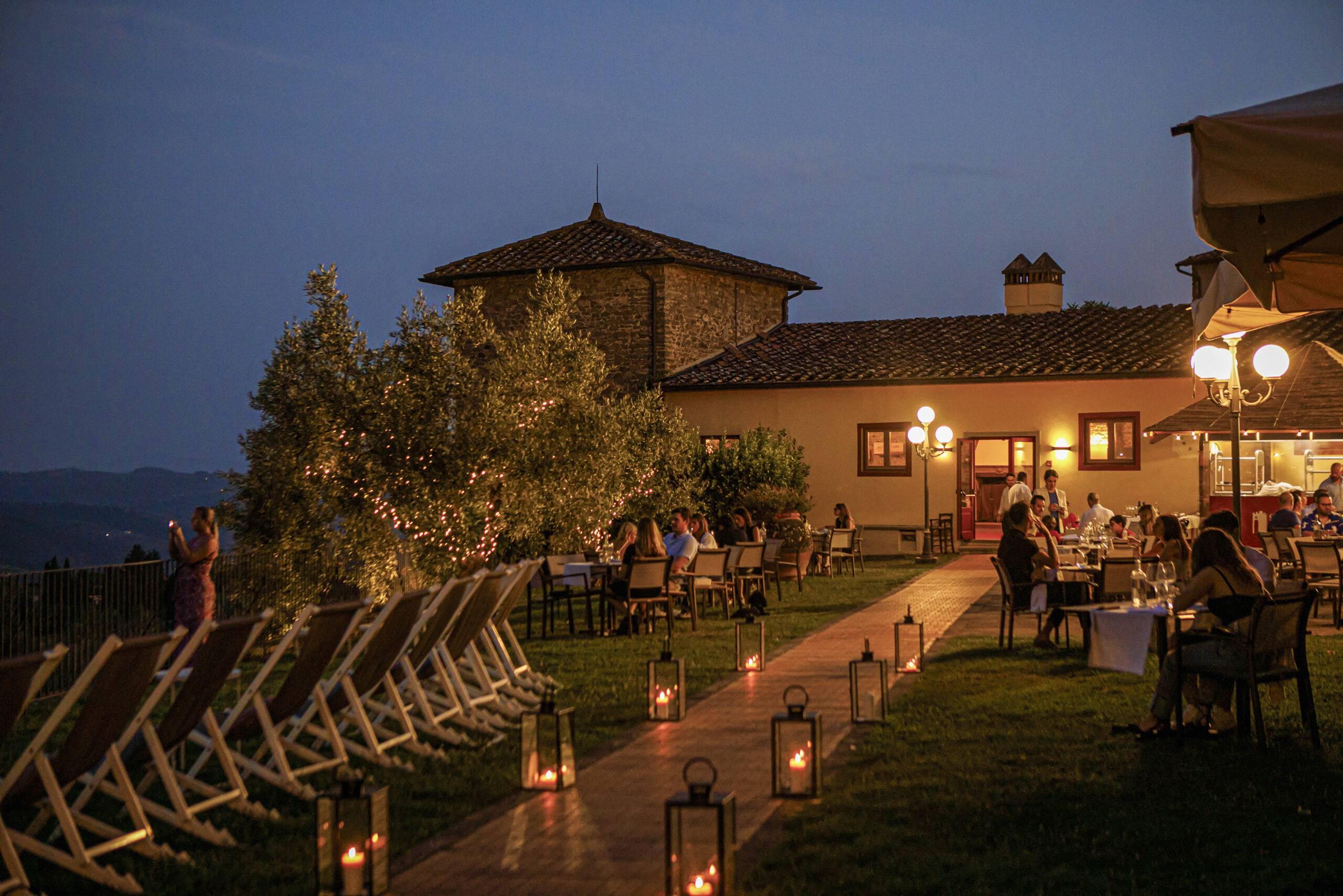 Evening outdoor view of the villa with a pathway lined by lanterns, tables set up on the lawn where people are dining, and warm lights on.