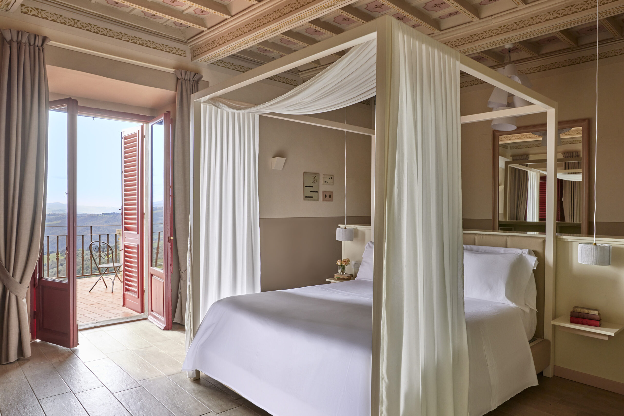 Interior of a bedroom with white bed and curtains around it, tile floors and a red door opening out onto a terrace.