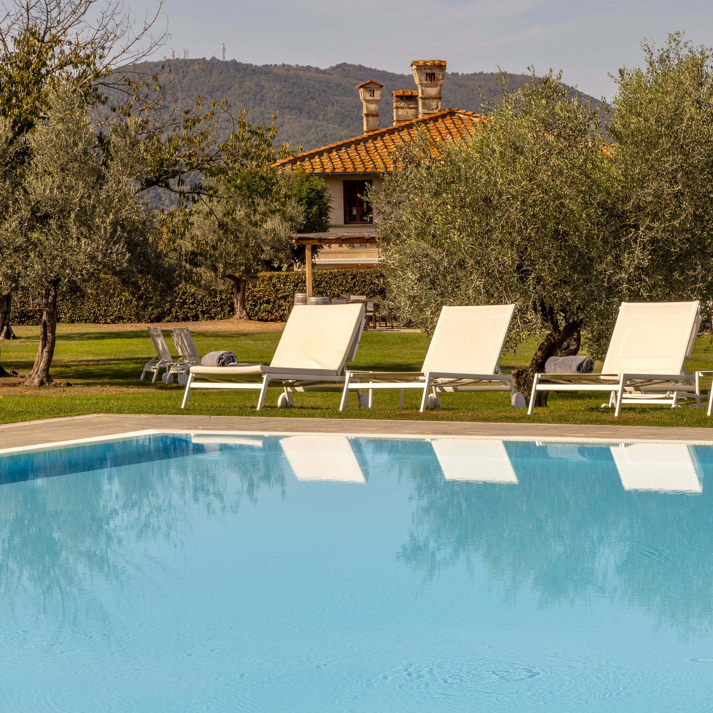 Three lounge chairs set up beside a turquoise swimming pool, with lawns, trees and the villa in the background.