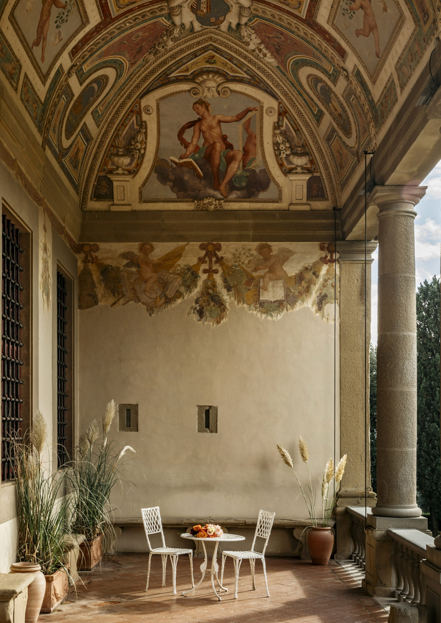 Terrace with a single table and two chairs sitting within a columned area with murals painted on the ceiling.