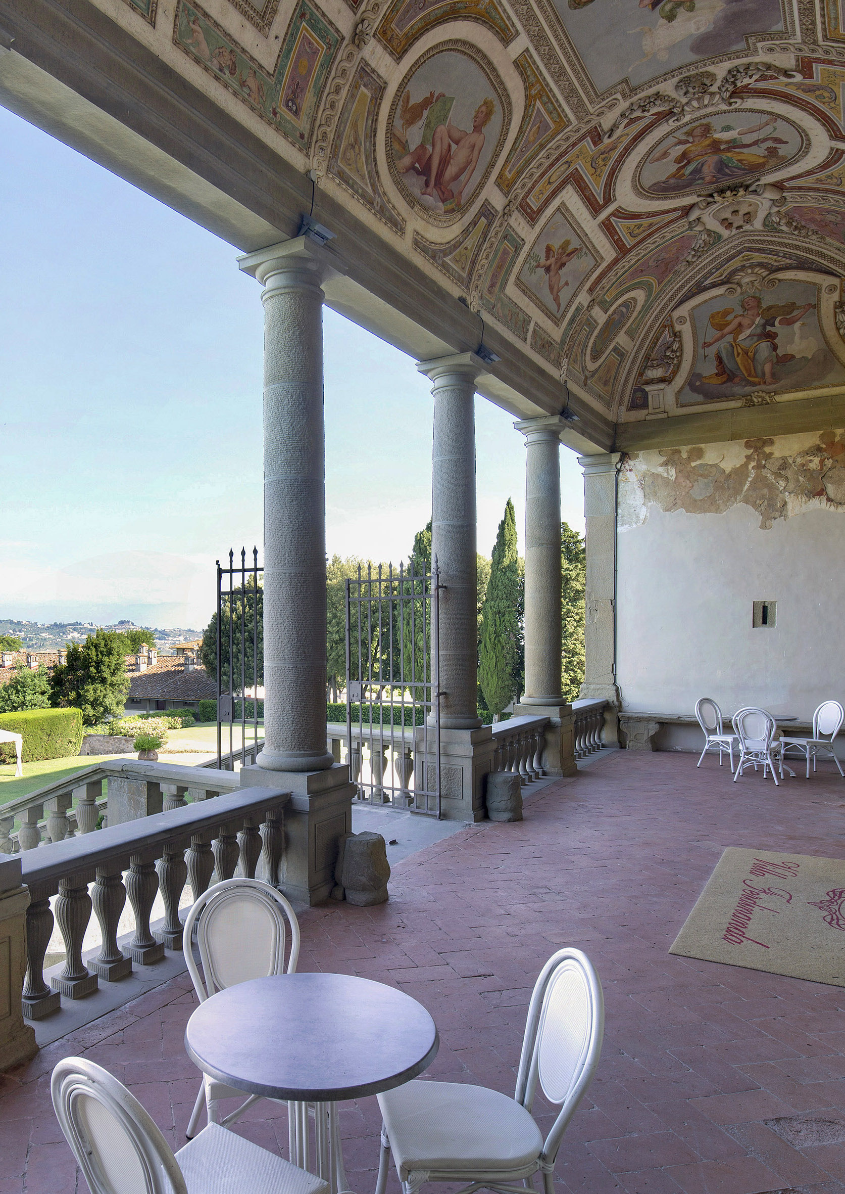 Terrace with a single table and two chairs sitting within a columned area with murals painted on the ceiling and a view out onto the countryside.