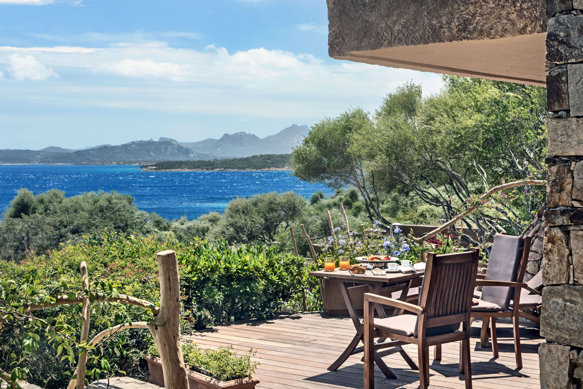 View of a wooden terrace with two deckchairs and a table overlooking greenery and the sea.
