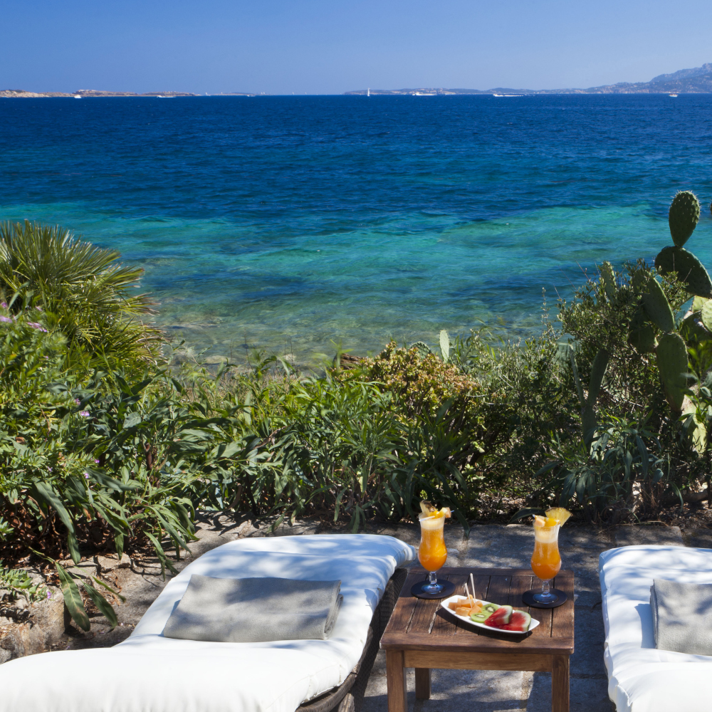 Two white lounge chairs with a table containing fruity drinks in between, overlooking greenery and a turquoise sea view.