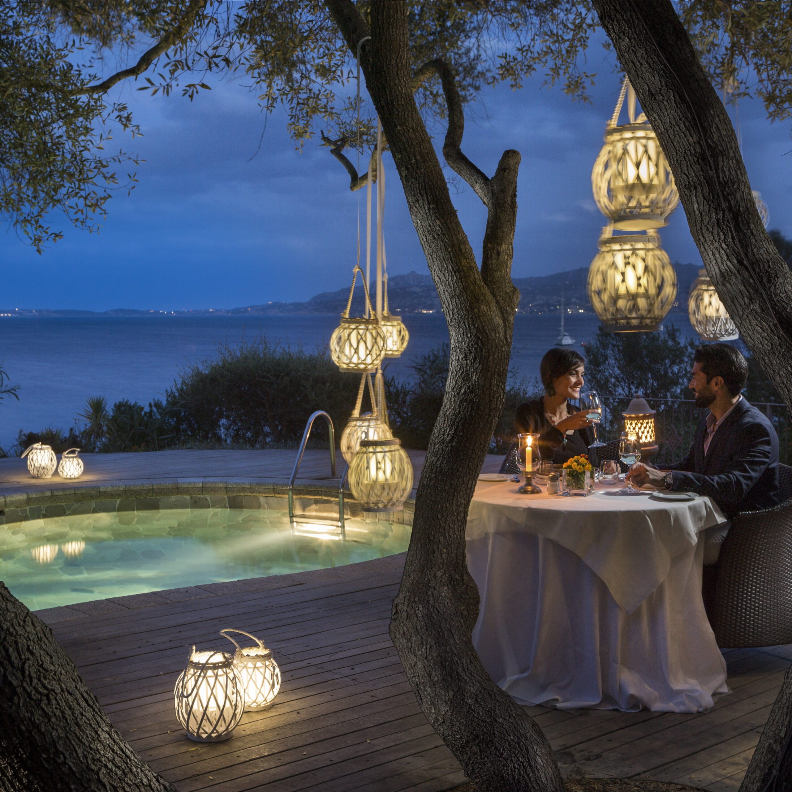 Evening scene of a people seated at an outdoor dining table surrounded by warmly lit lanterns, the pool and sea beyond.