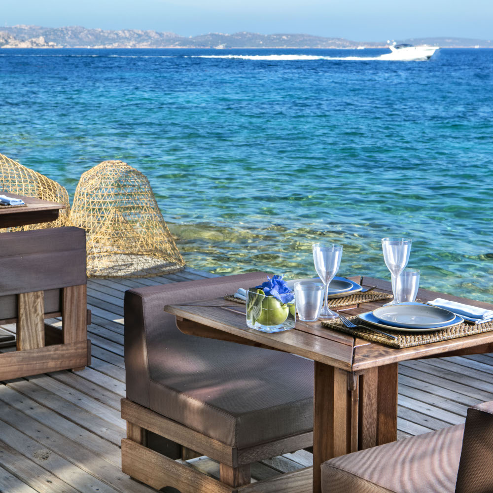 Small low brown tables and chairs set with wine glasses and plates alongside the sea.