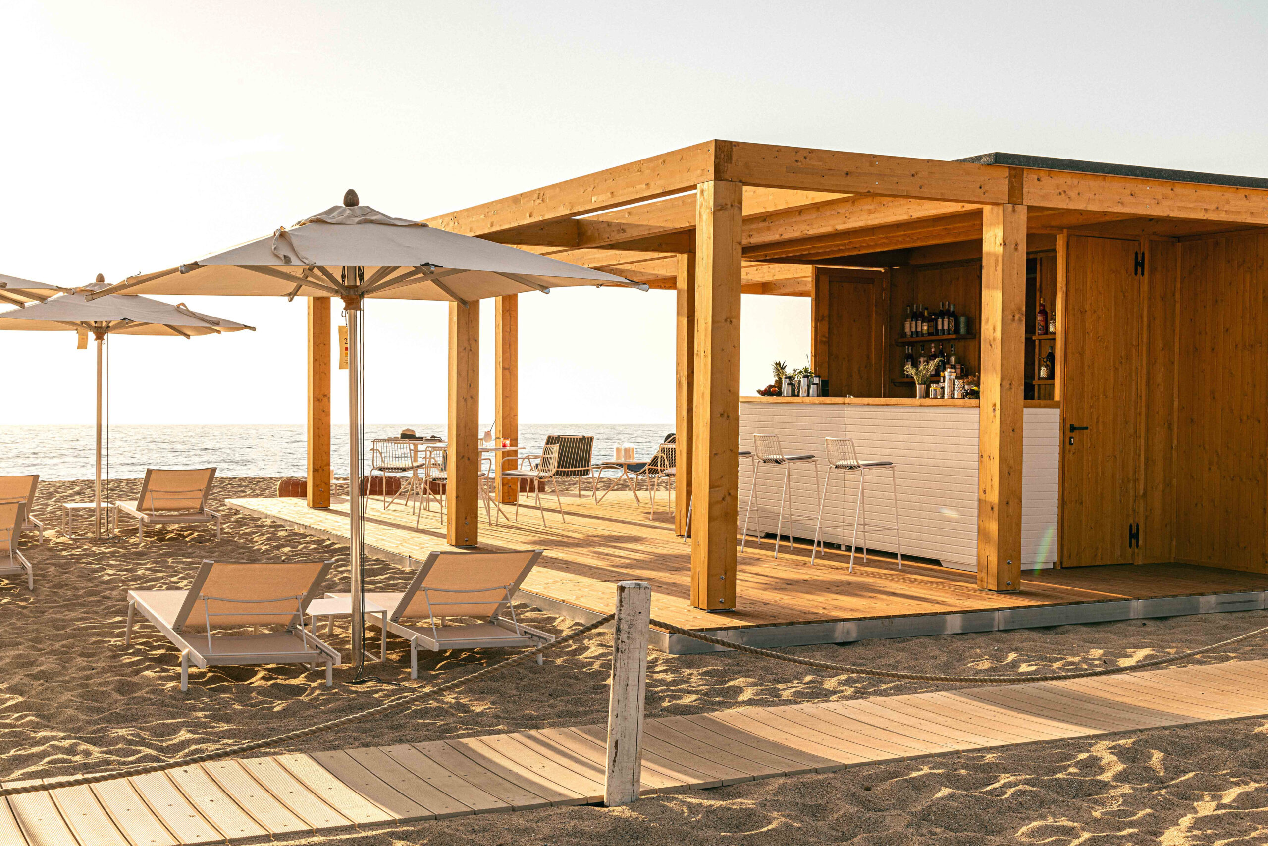 Outdoor wooden bar structure set on the sand with a few umbrellas and lounge chairs.