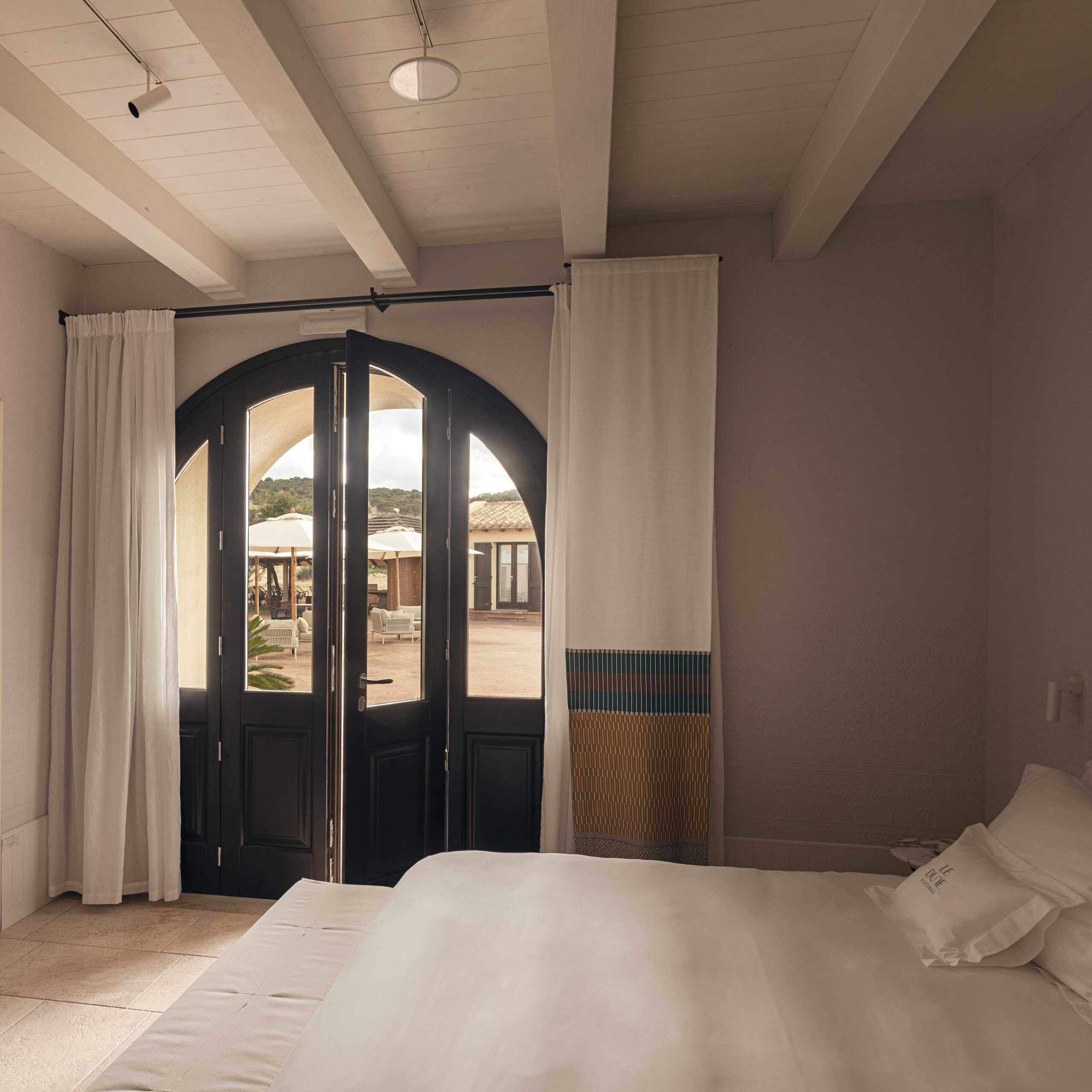 Interior of a bedroom with a white bed in the foreground and an arched doorway framed by curtains leading outside.