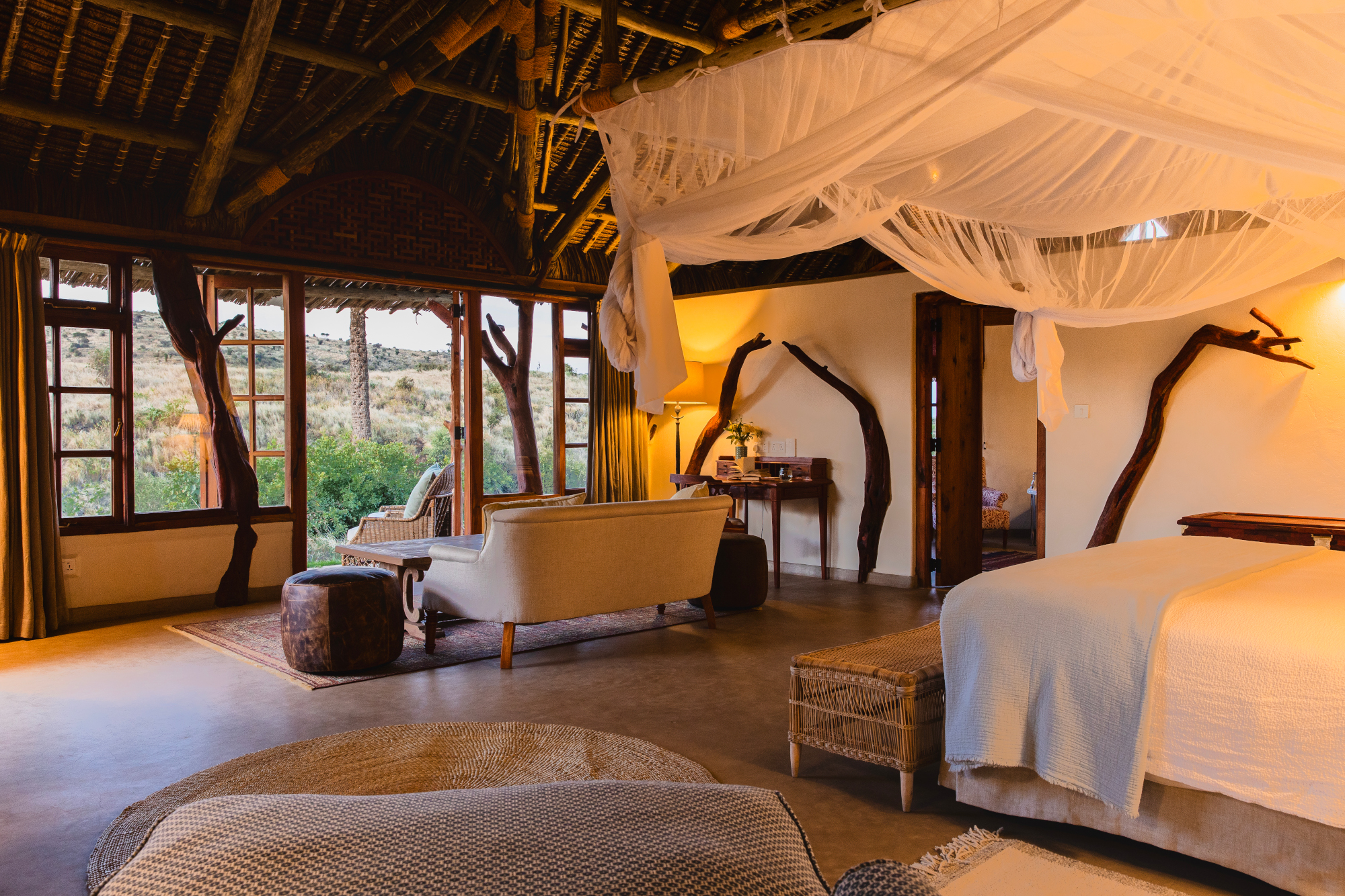 A safari bedroom with a bed with white mosquito netting, a sofa and wooden thatch ceiling and windows looking out onto the bush.