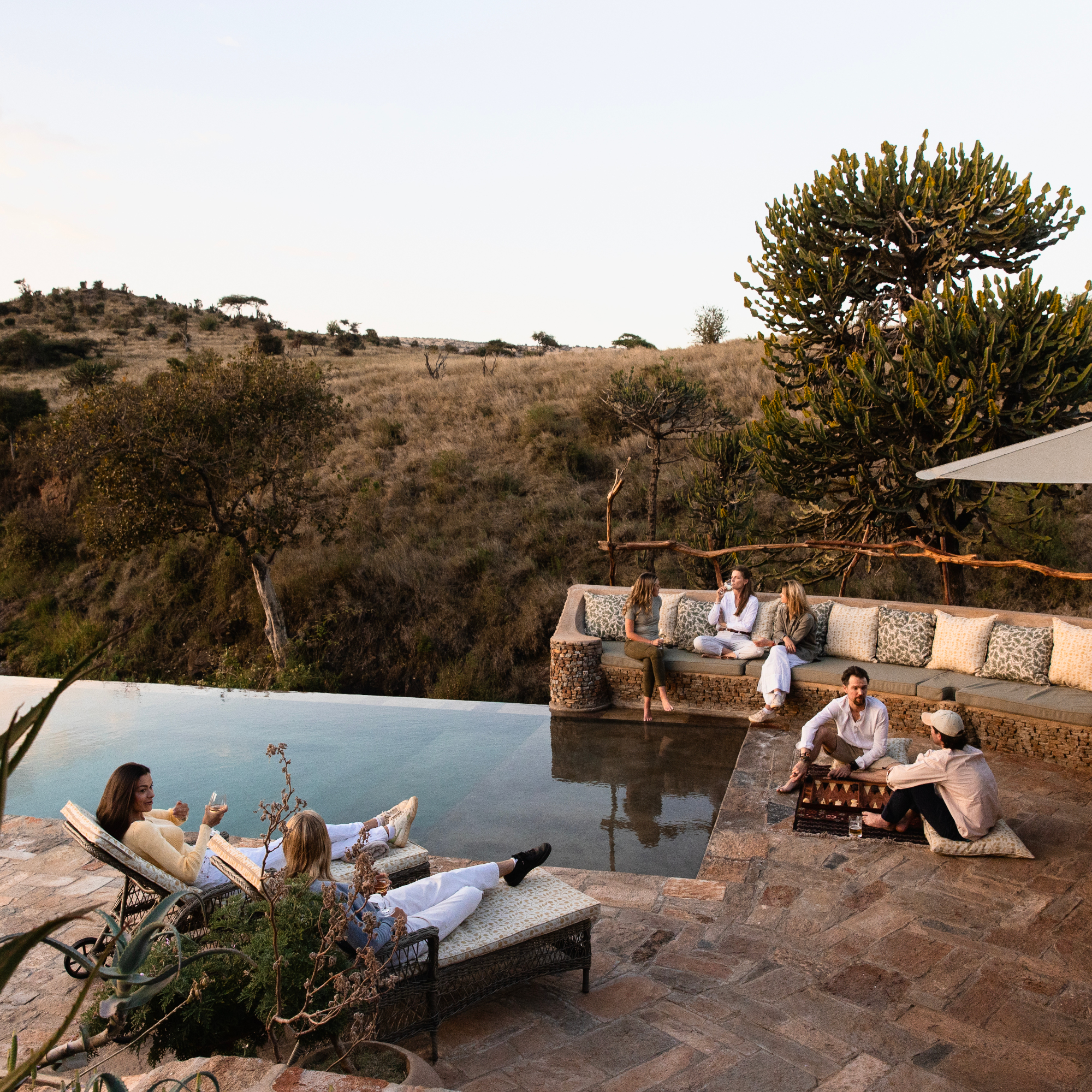 Several people sitting on lounge chairs around an infinity pool overlooking the Kenyan bushland beyond.