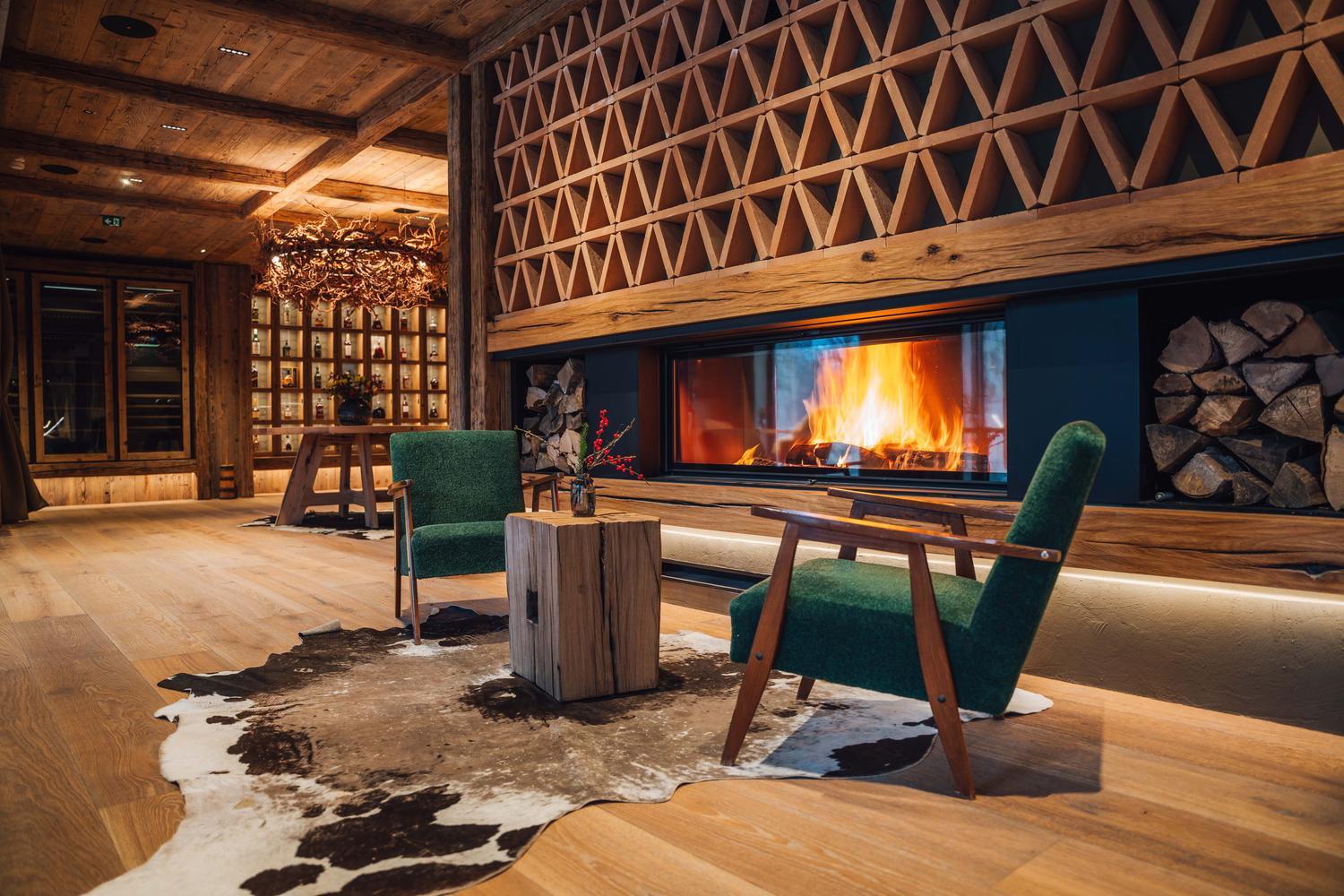 Stylish lounge with a burning fireplace, two green chairs on a cowhide rug, and a geometric wood-carved wall.