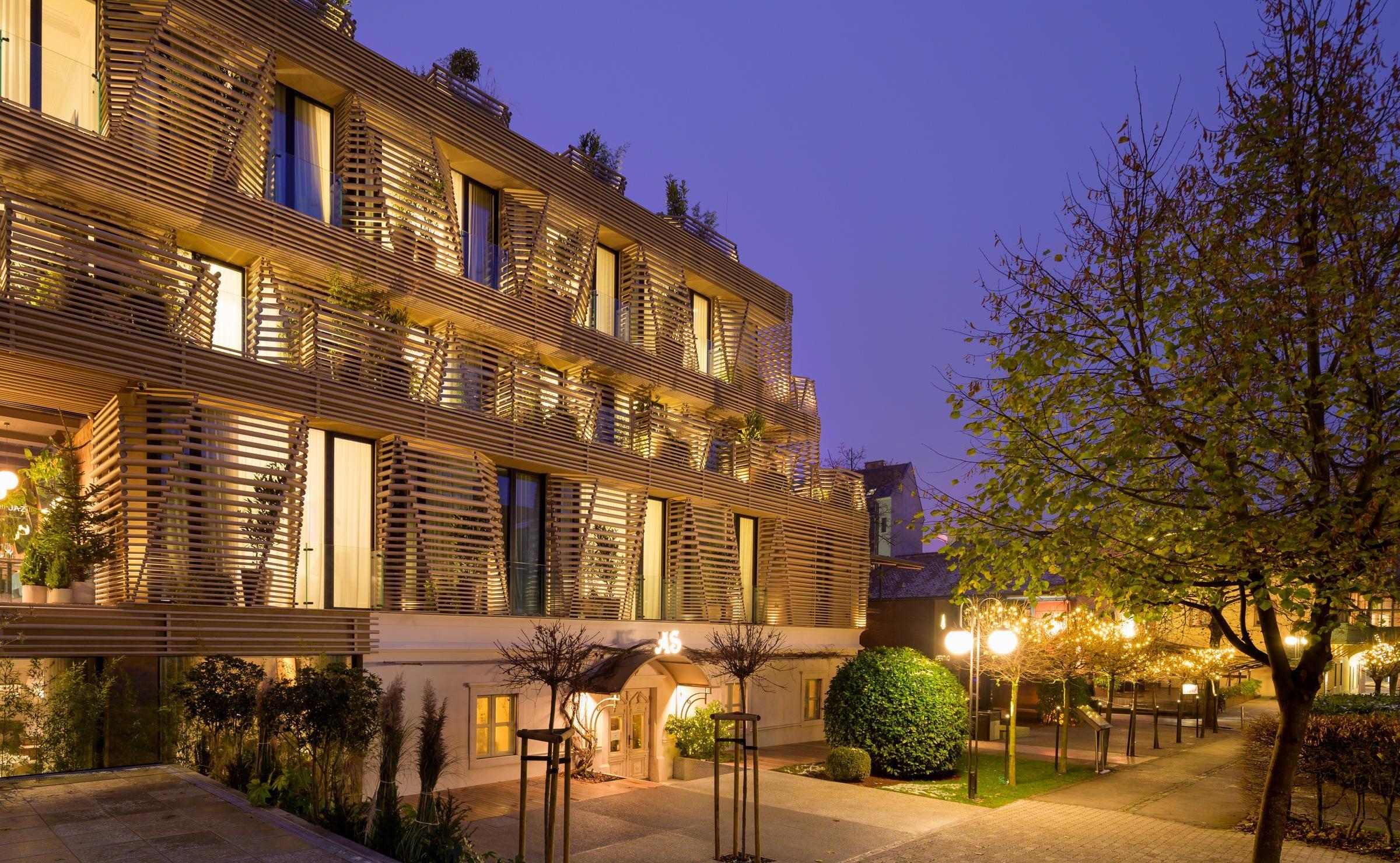Night exterior of AS Boutique Hotel in Ljubljana with illuminated wooden slats and surrounding green trees.