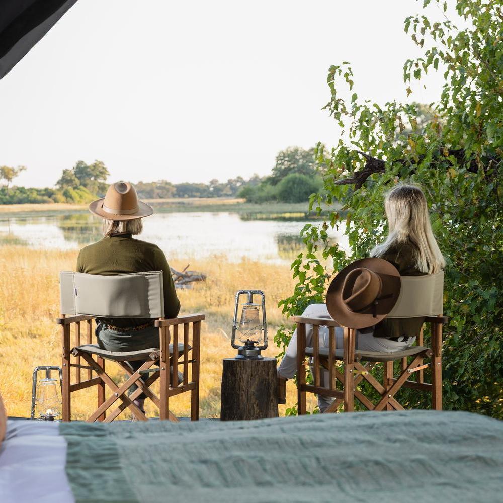 Back view of two people in safari hats sitting in chairs looking out at a calm waterhole and trees.