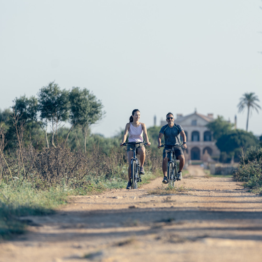 Two people riding bicycles on a dirt path near Son Vell in Balearic Islands Spain with a manor house behind them.
