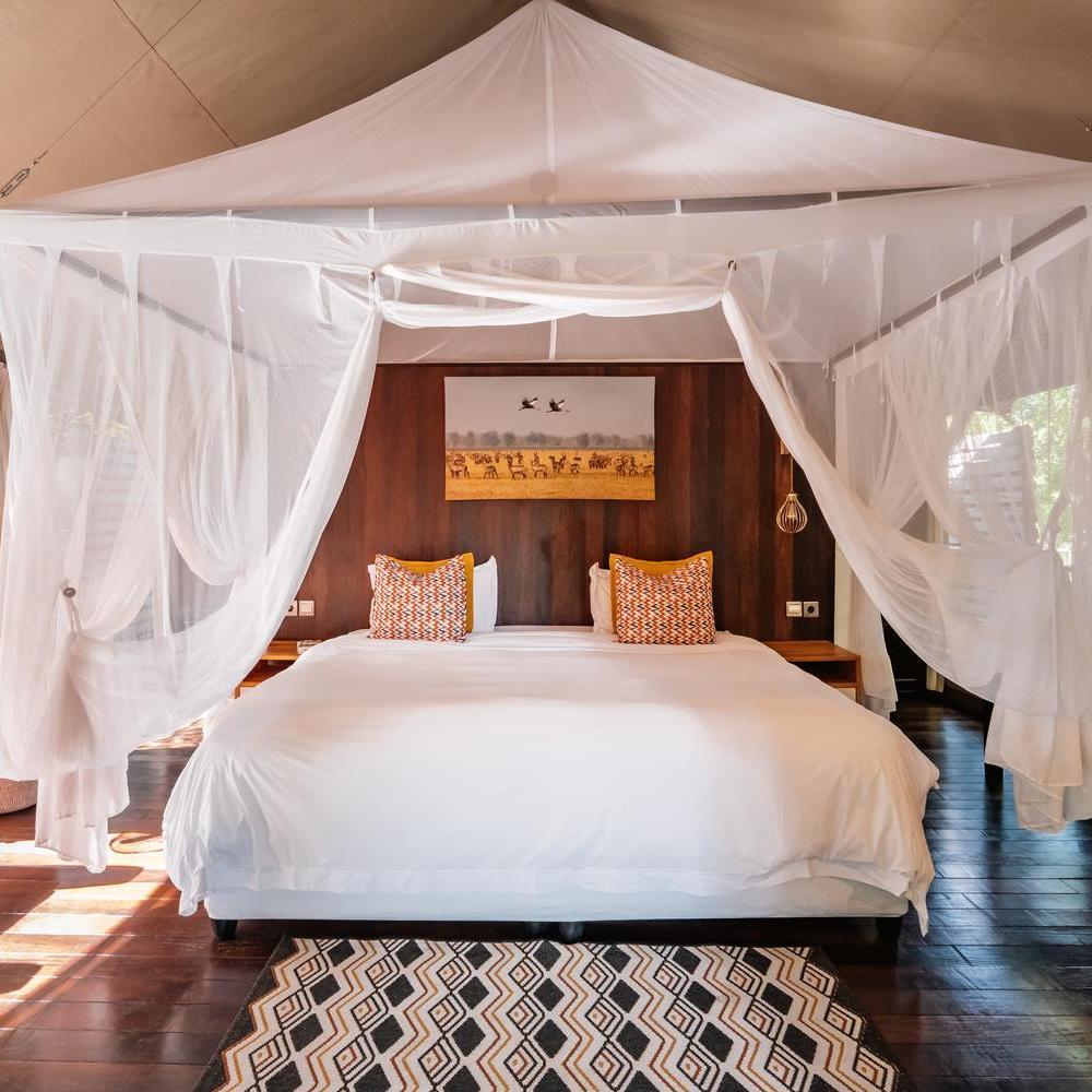 A king-sized bed with a white mosquito net canopy and decorative pillows inside a wooden-walled safari tent.