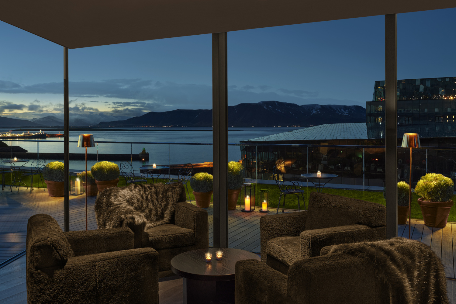 Interior of a bar table with dark grey sofa chairs in a corner with floor-to-ceiling windows looking out over a harbour a dusk.