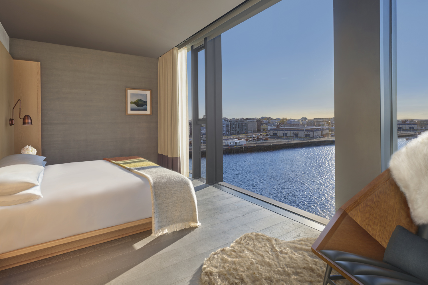 Bedroom with grey walls and a bed with white and tan sheets facing a floor-to-ceiling window overlooking the harbour.