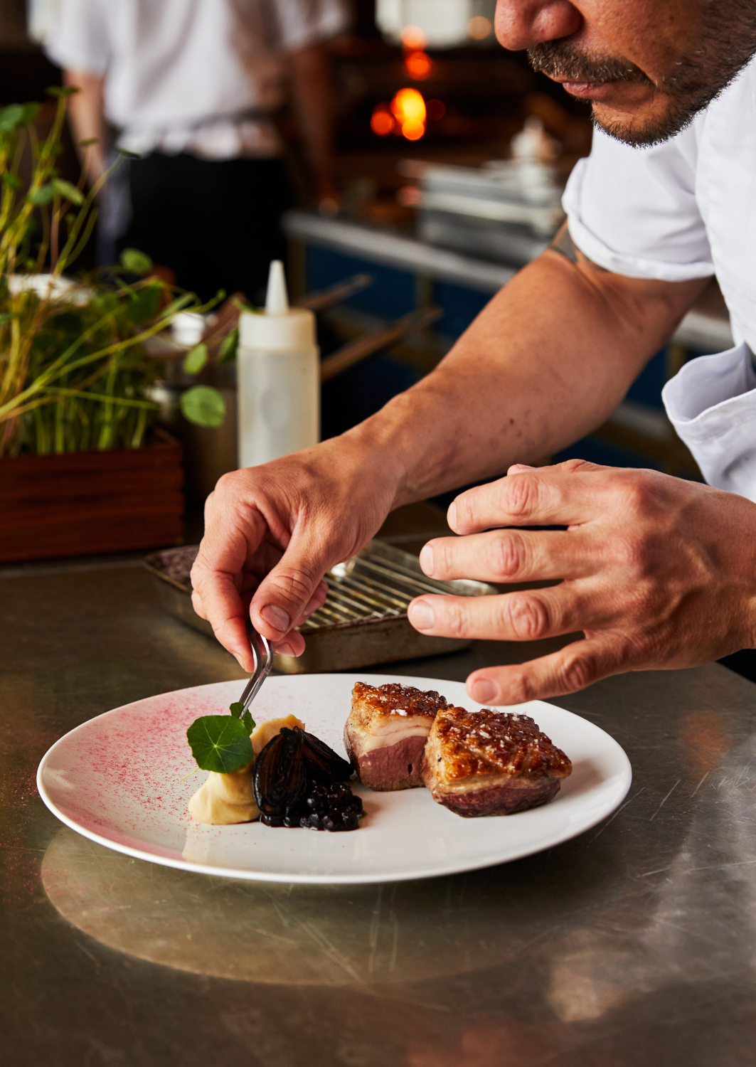 A chef's hands plating food onto a plane white plate with a candle burning in the background.
