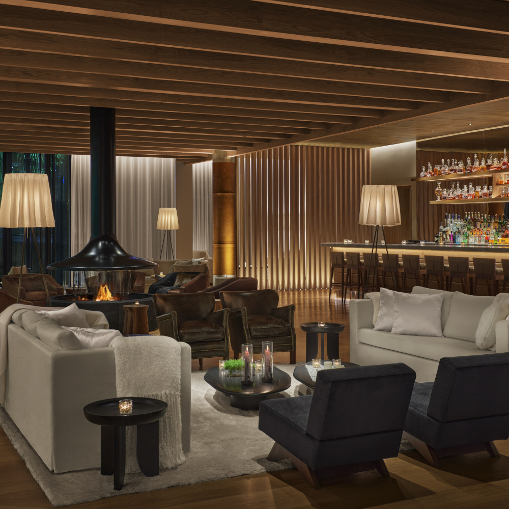 Interior of a bar with white sofa and black chairs, a light wood ceiling and warm lamps, a bar in the background and in the back an open wood stove.
