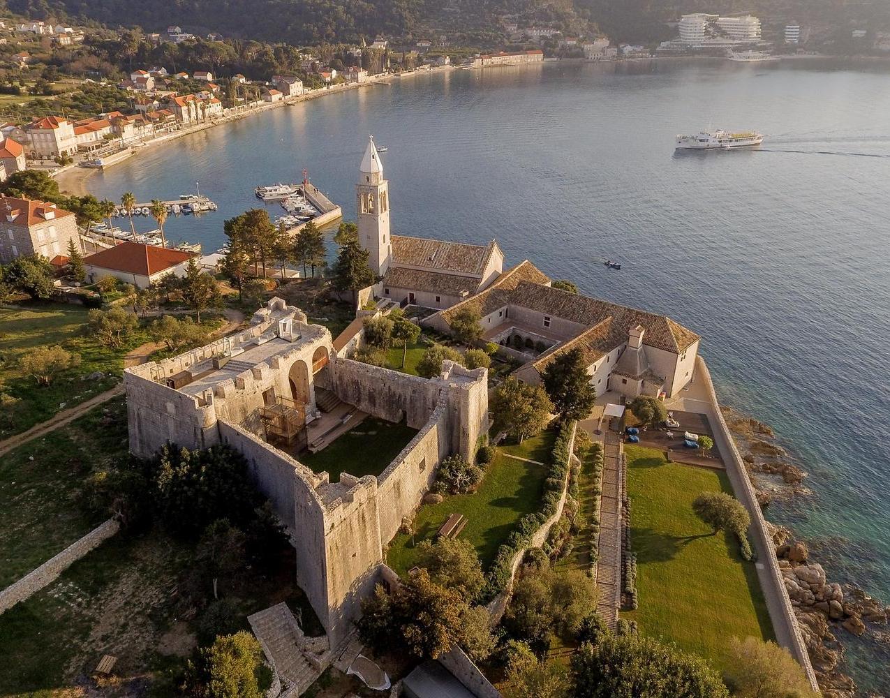 High-angle aerial view of a stone monastery and fortress complex on the coast of Lopud island near a calm bay.