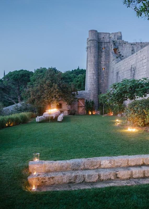 A long dining table set with candles in a grassy garden next to a tall, illuminated stone fortress tower.